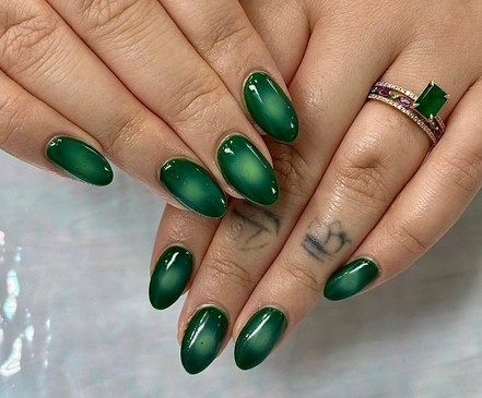Hand with green manicured nails, rings, and a tattoo of an anchor on the finger, against a neutral background.