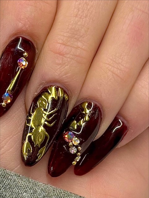 Close-up of five fingers with dark red and gold nail art, decorated with rhinestones and intricate designs.