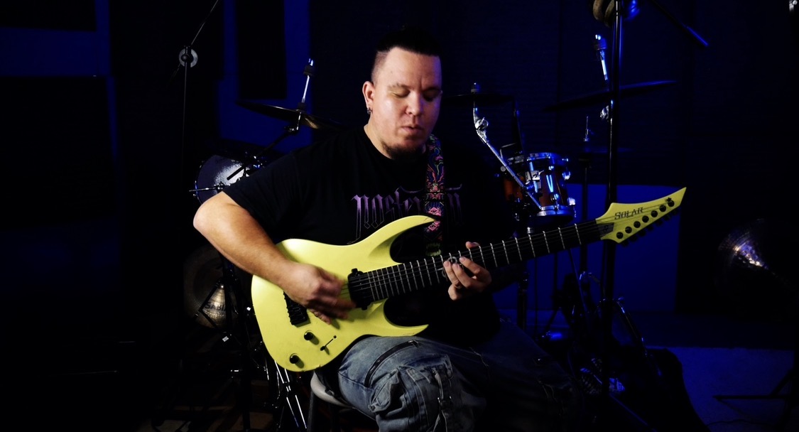 Person playing yellow electric guitar on stage with drums in background.