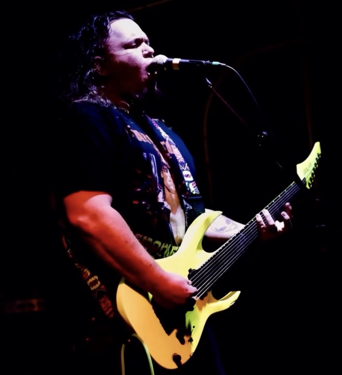 Person singing into microphone while playing an electric guitar on stage, dark background, focused lighting.