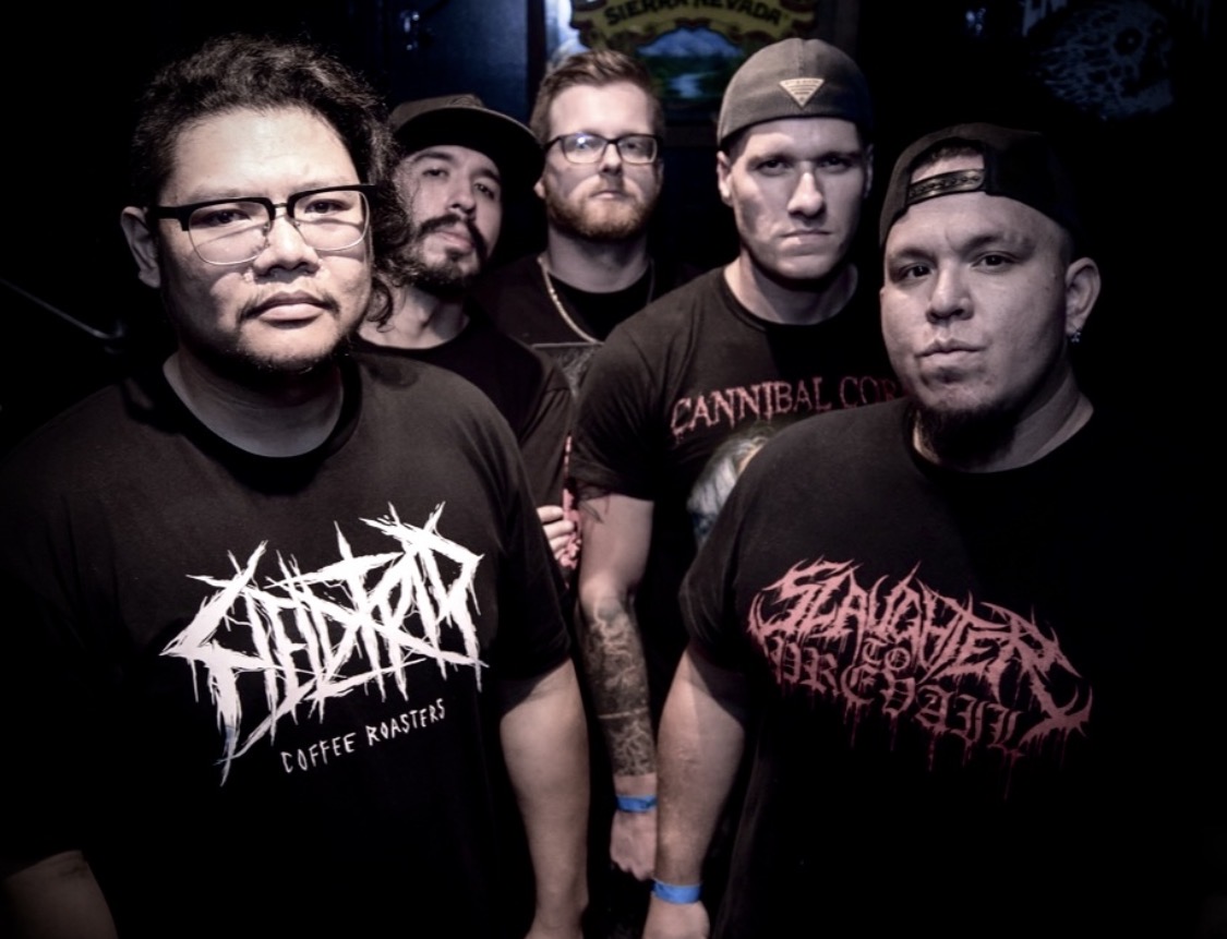 Group of five men with dark clothing and graphic t-shirts, standing indoors with dark background.