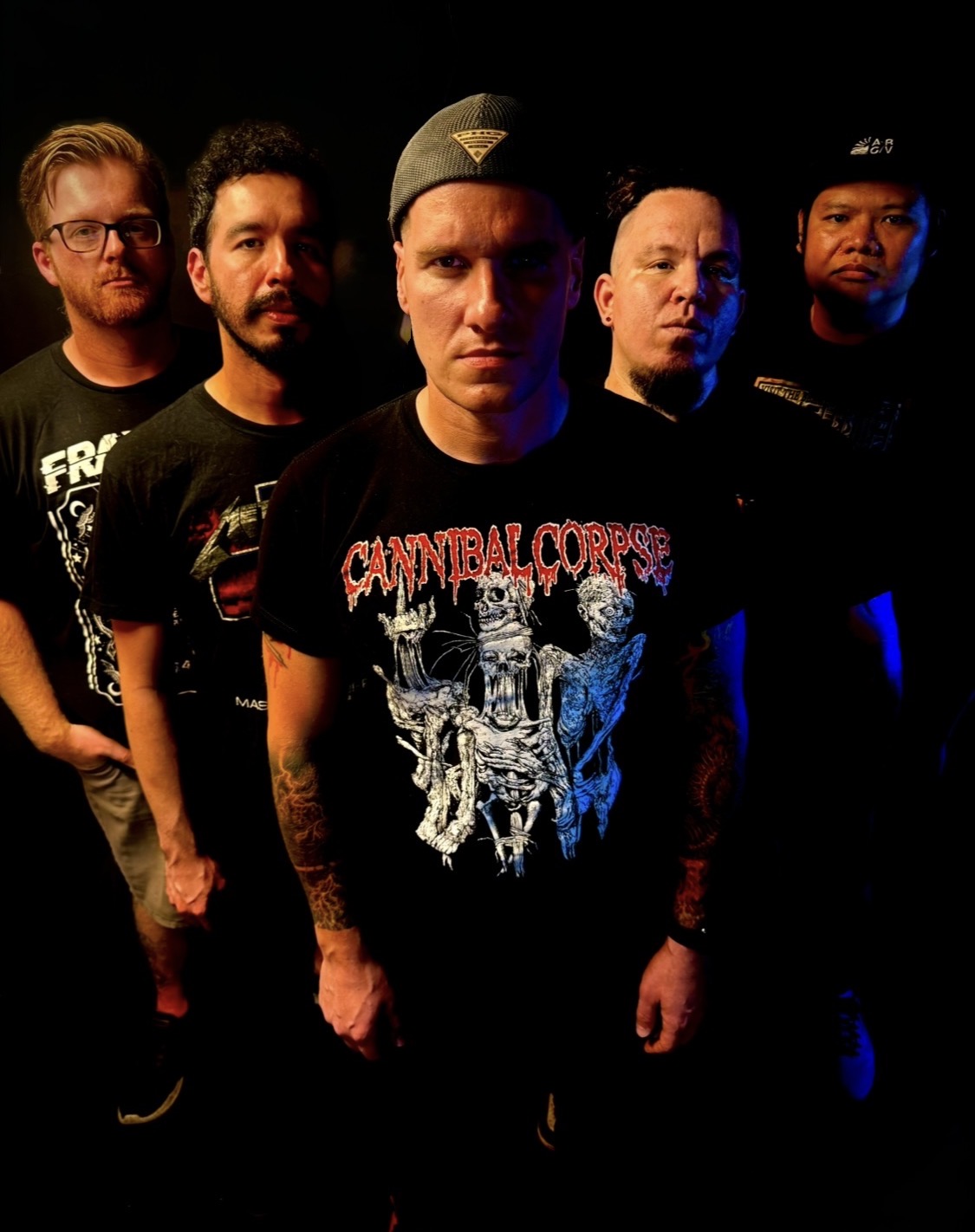 Five men standing in a row against a dark background, some with serious expressions, wearing casual clothing and hats.