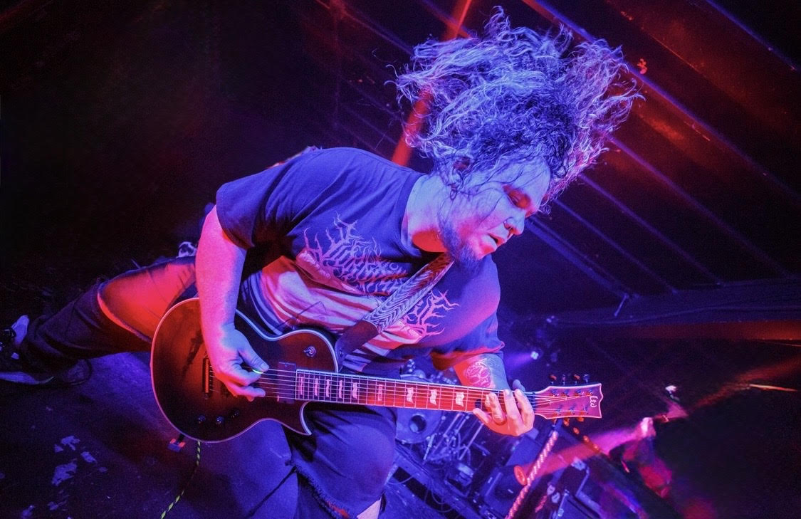 Person playing guitar with long hair flying, colorful stage lights, energetic performance, dark background.