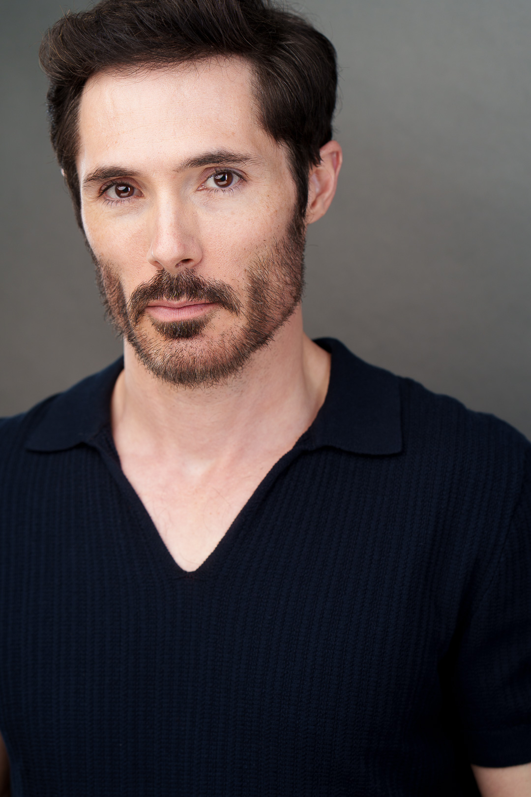Man with dark hair and beard wearing a dark V-neck shirt, looking at the camera against a plain background.