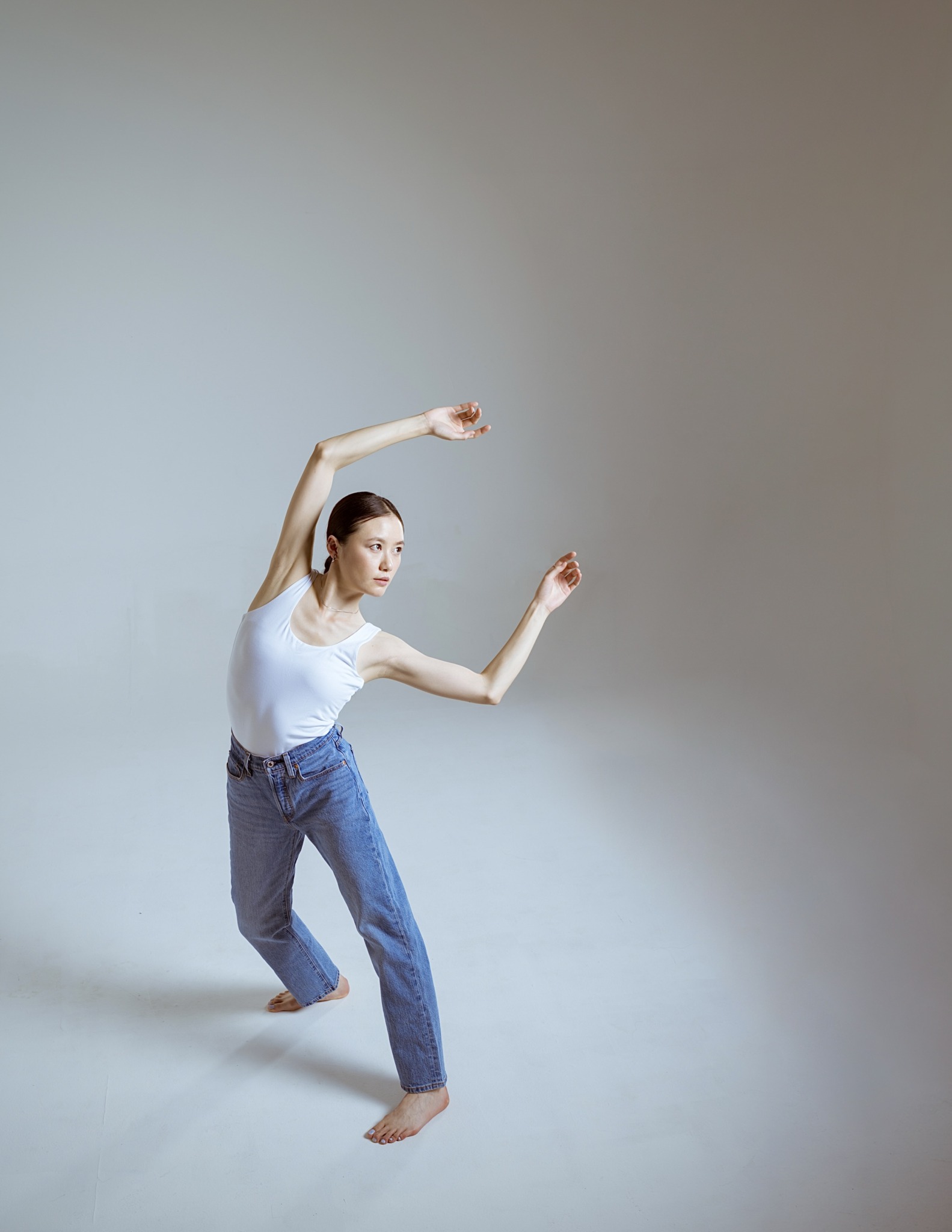 Person in a white top and jeans poses with arms raised and bent, standing on one foot against a plain background.