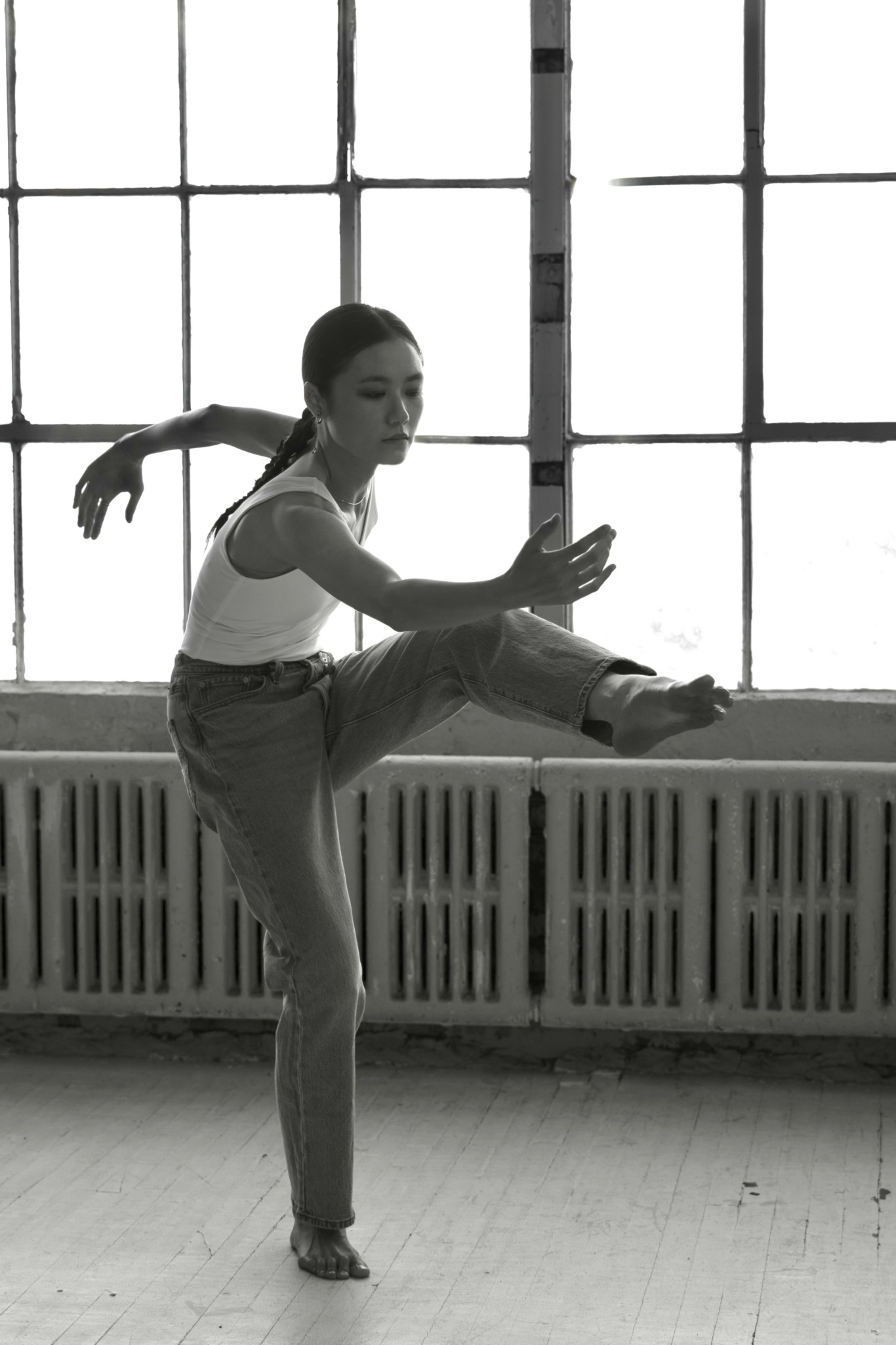 A woman practicing ballet or dance in front of large window panes, balancing on one leg with arms extended.