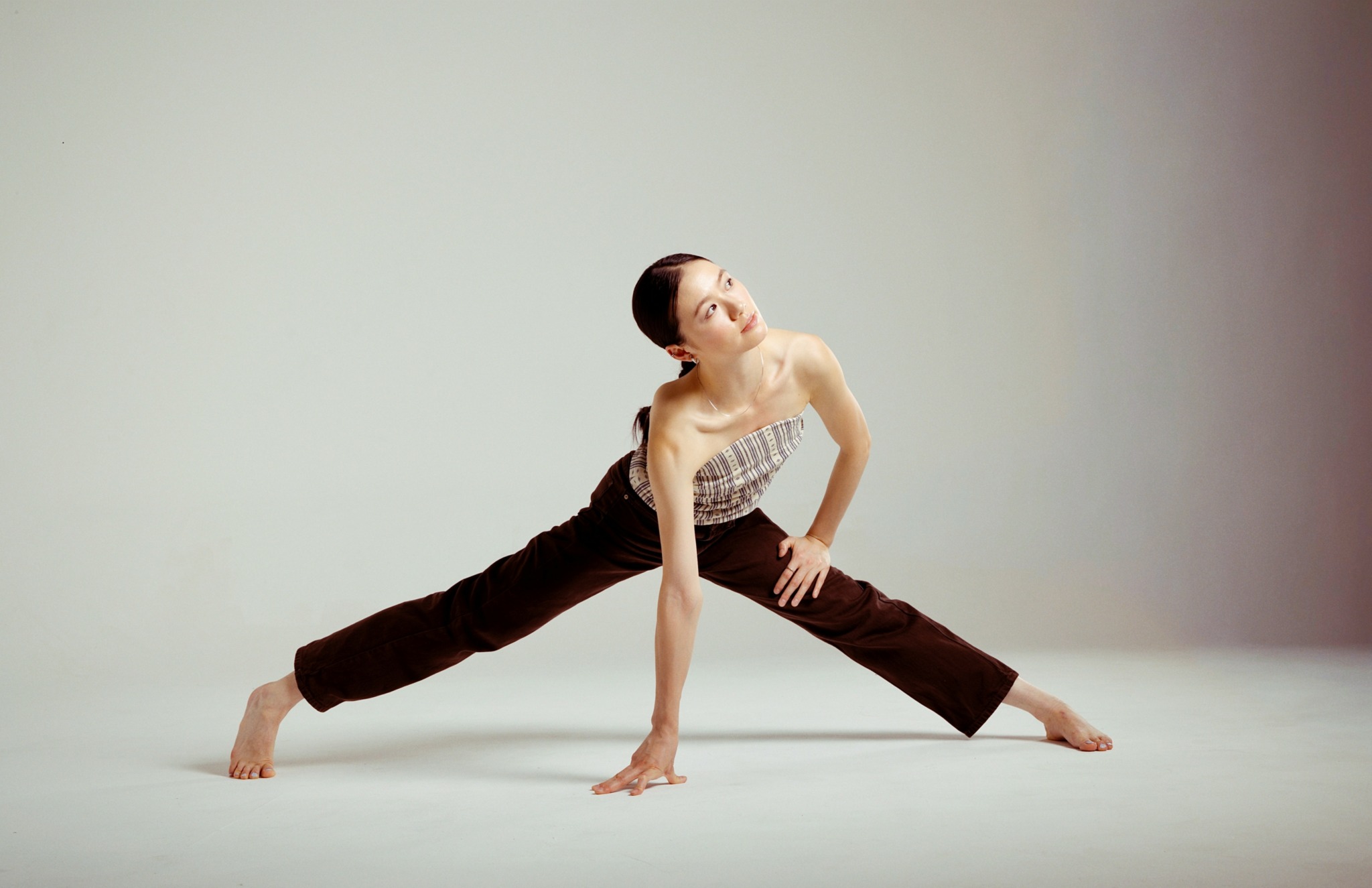A woman in a dance pose stretches on a plain background, with one hand on the floor and the other arm extended upward.