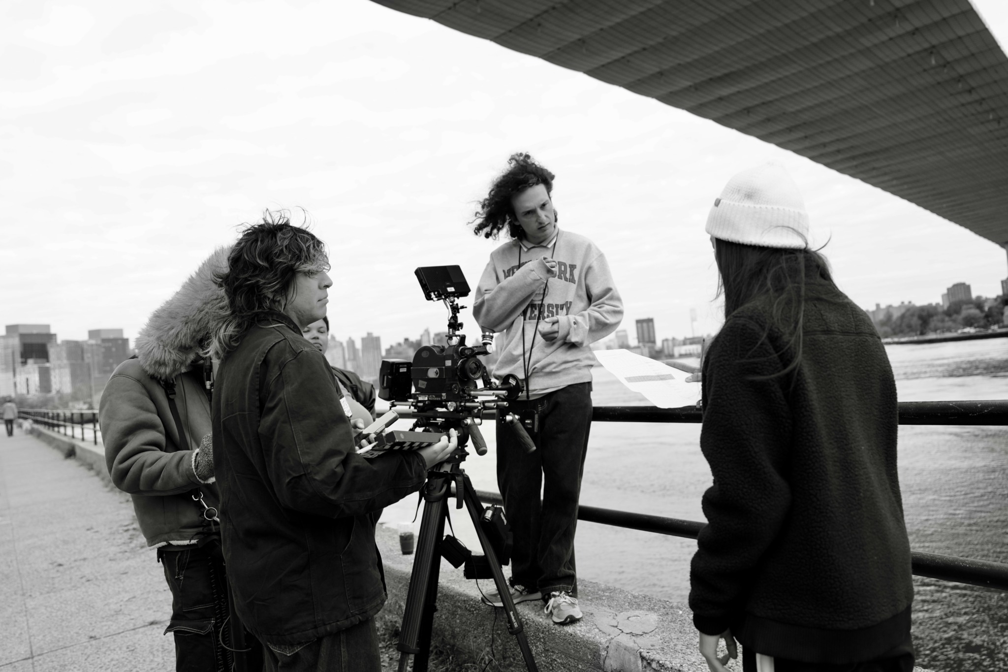 Four people, one with a camera, are outdoors near a river with a city skyline in the background.