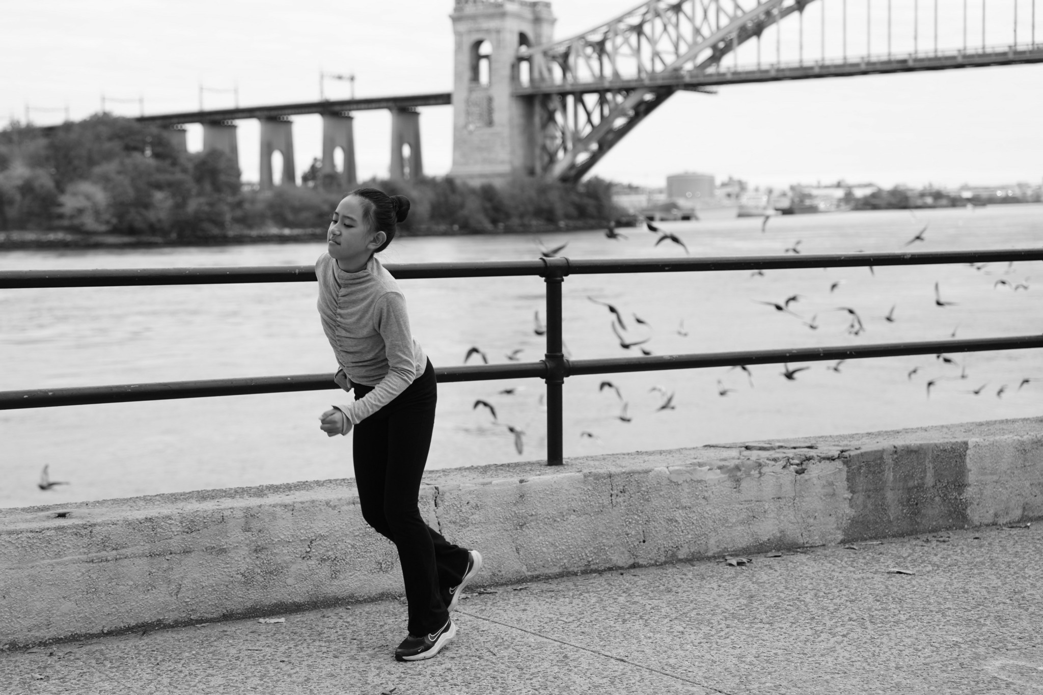 Woman jogging along a riverside with a bridge in the background, birds flying nearby, black and white photo.