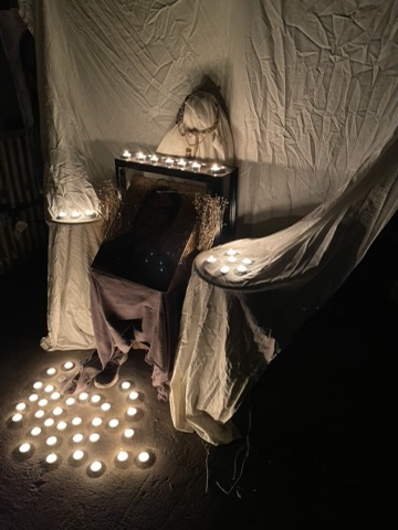 A dimly lit scene with candles on a table, a person sitting, and fabric draped around, creating a cozy atmosphere.