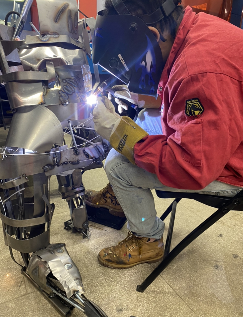 Person in red jacket welding a metallic robotic arm, wearing a helmet and gloves, seated on a black chair.