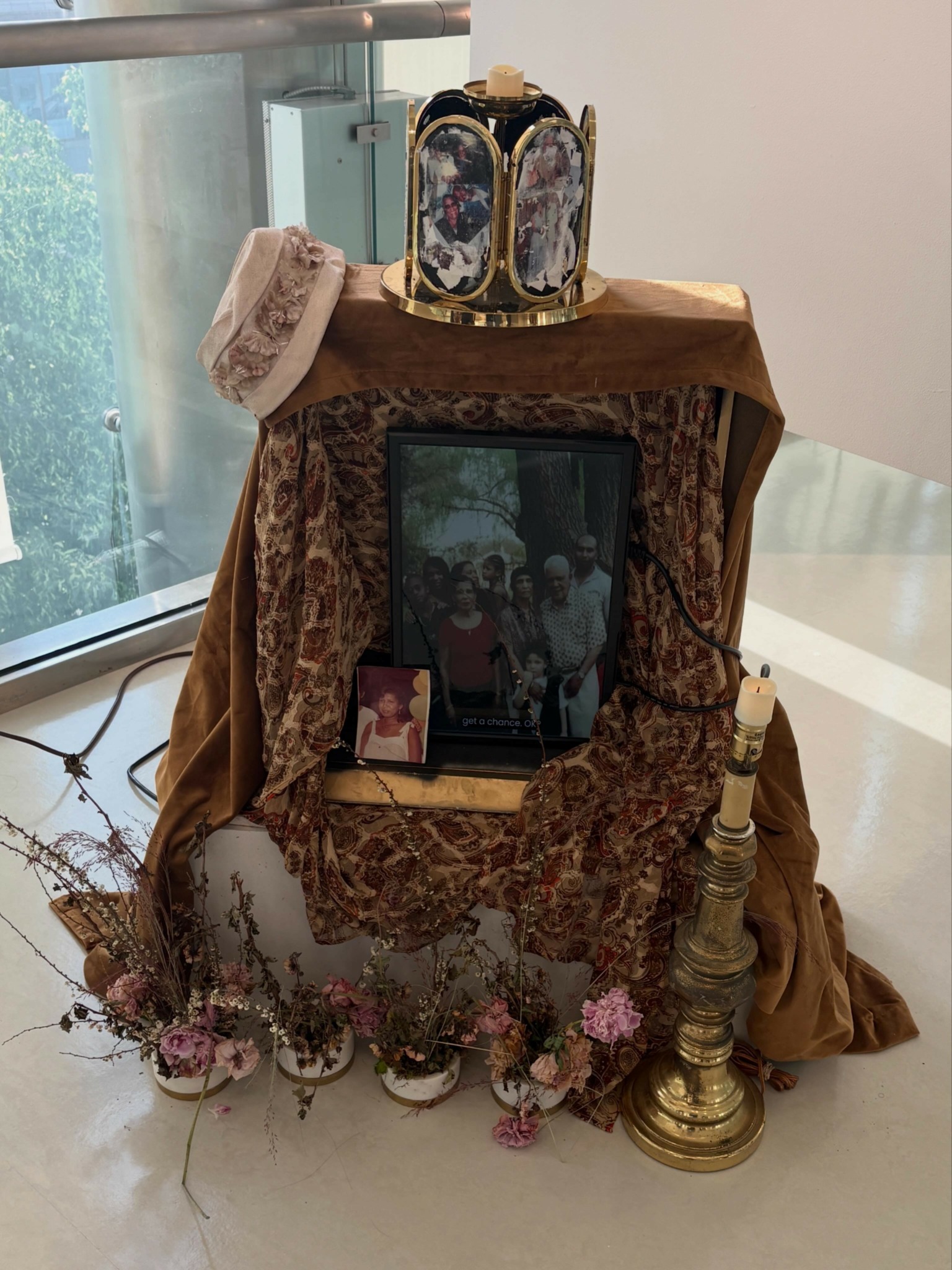 Display with framed photos, flowers, and decorative items on a table near a window, with a cloth backdrop.