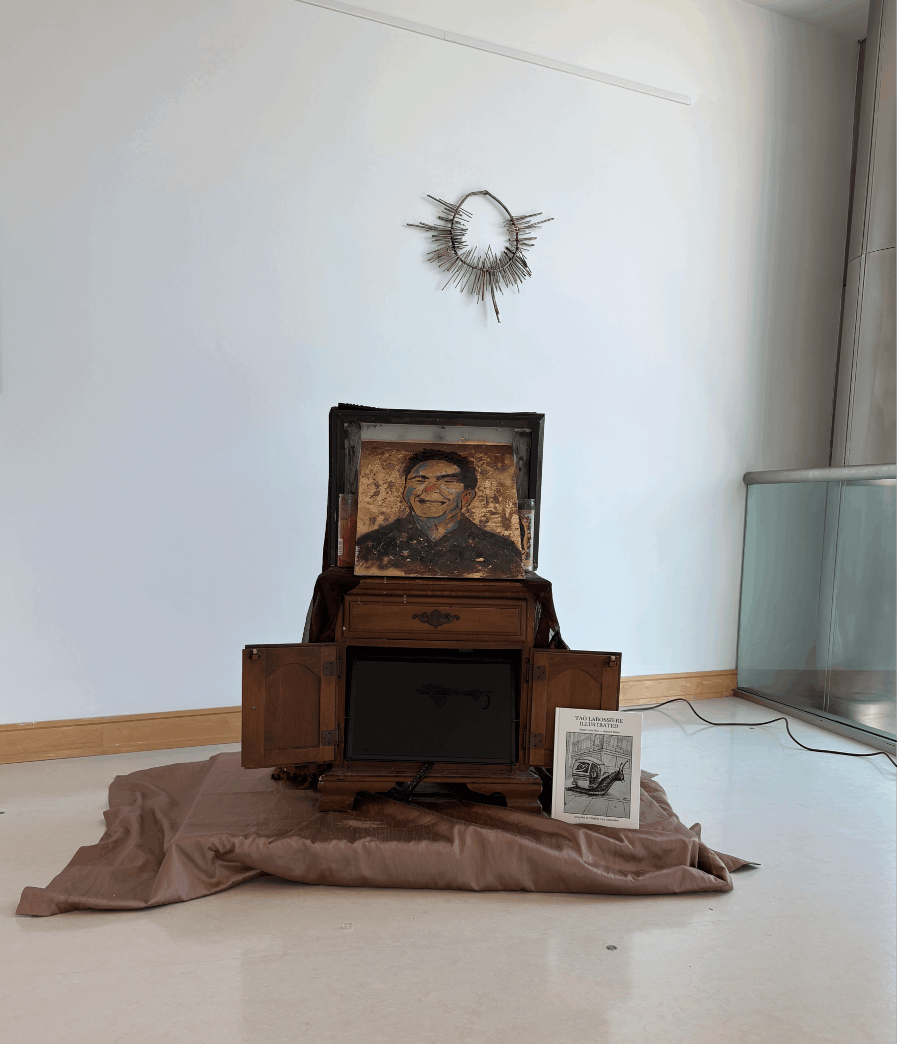 A framed portrait of a person is placed inside an open wooden box on a cloth-covered surface, with a small black object and a paper nearby. A decorative circular wall hanging is above, and a glass display case is to the right.