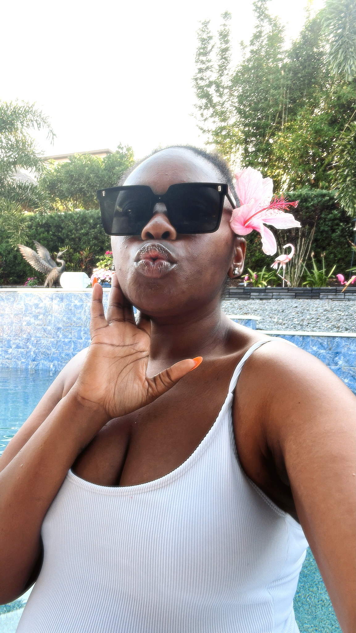 Woman with sunglasses and a pink flower in her hair, making a kiss face outdoors near a pool, greenery in background.