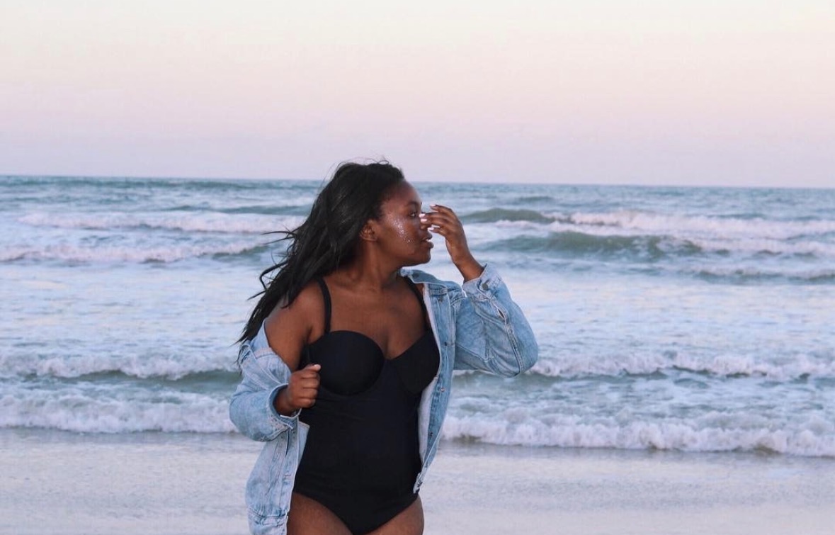 Woman in a black swimsuit and denim jacket standing on the beach, looking at the ocean, with waves in the background.