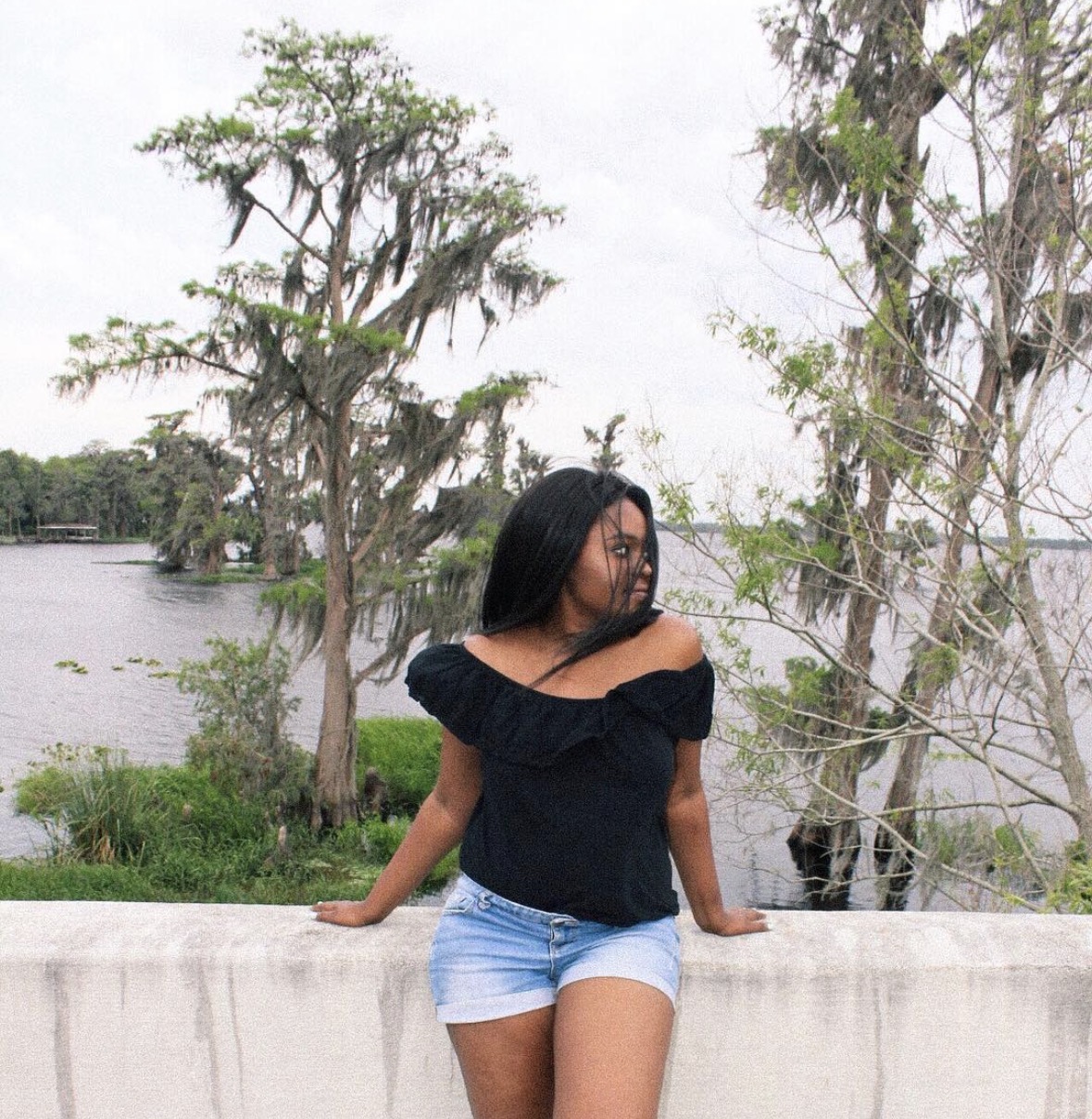 Woman with black hair in a black off-shoulder top and denim shorts leaning on a concrete barrier outdoors near a body of water with trees.