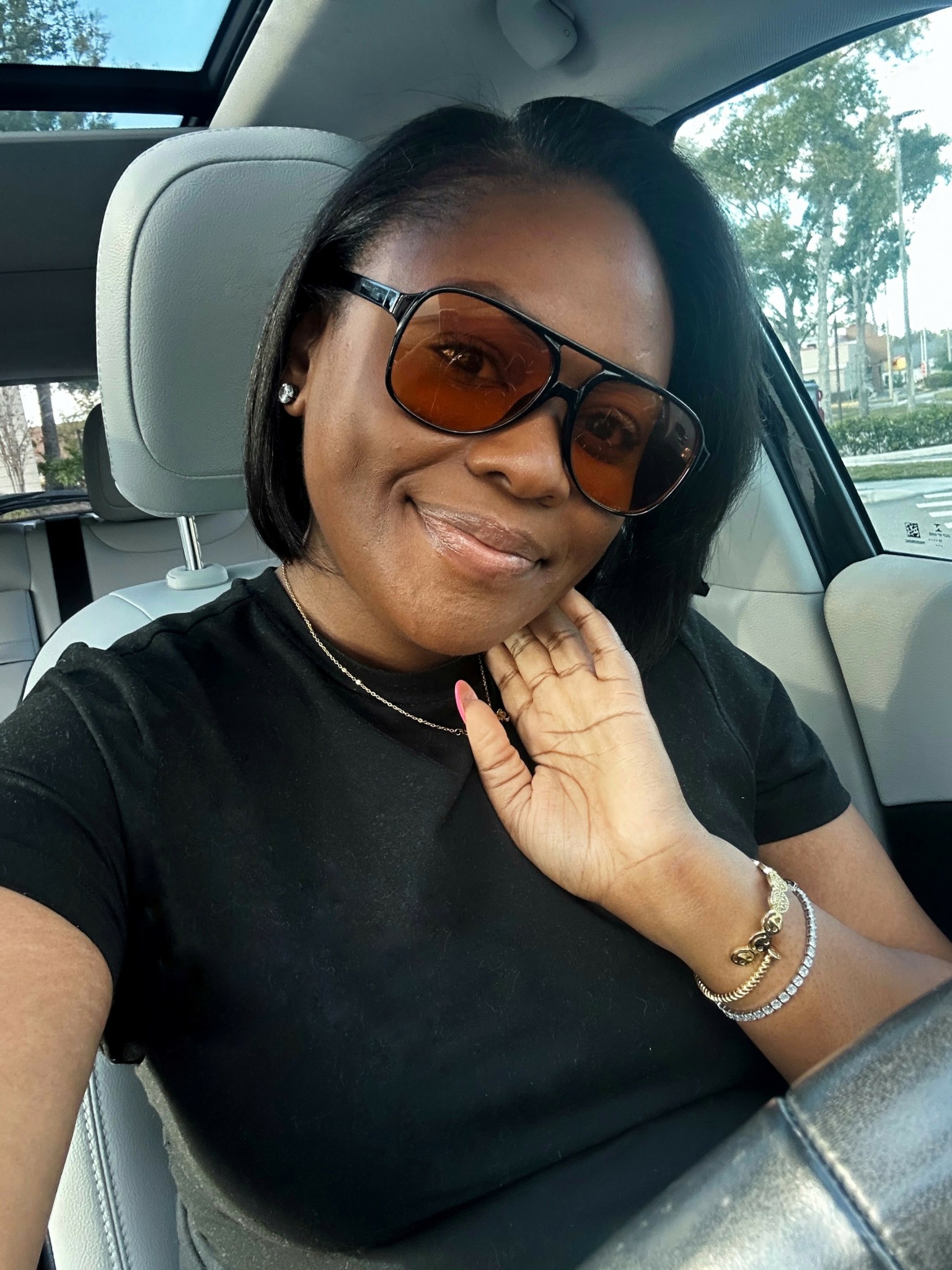 Woman with short black hair wearing sunglasses and a black shirt, sitting in a car, smiling, with trees outside the window.