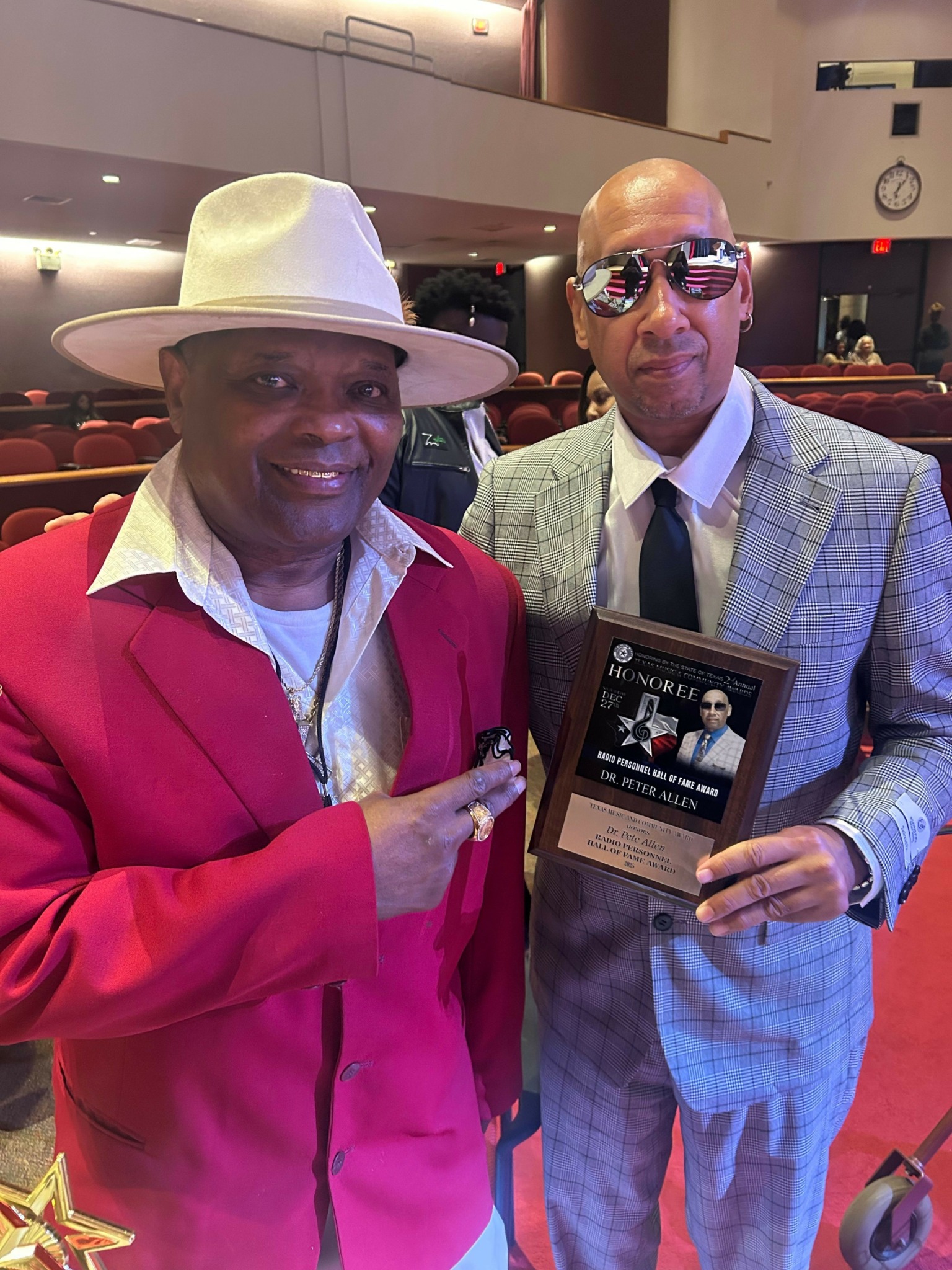 Two men, one in a pink jacket and white hat, and the other in a blue checkered suit, pose indoors with a plaque.