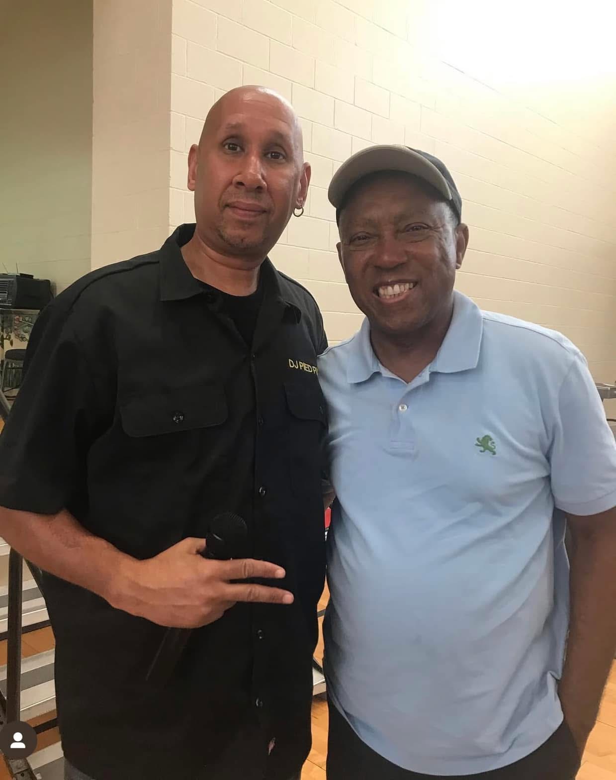 Two men standing indoors, one in a black shirt and the other in a light blue polo, smiling at the camera.