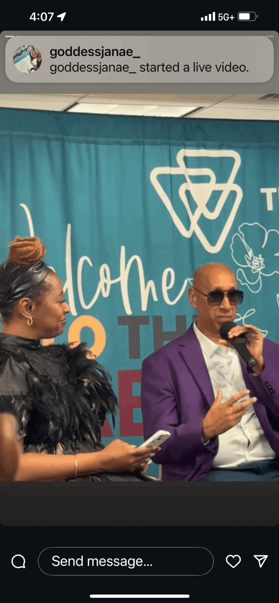 Two people on stage, one woman and one man, speaking into microphones, with a blue backdrop and a logo, at an event.