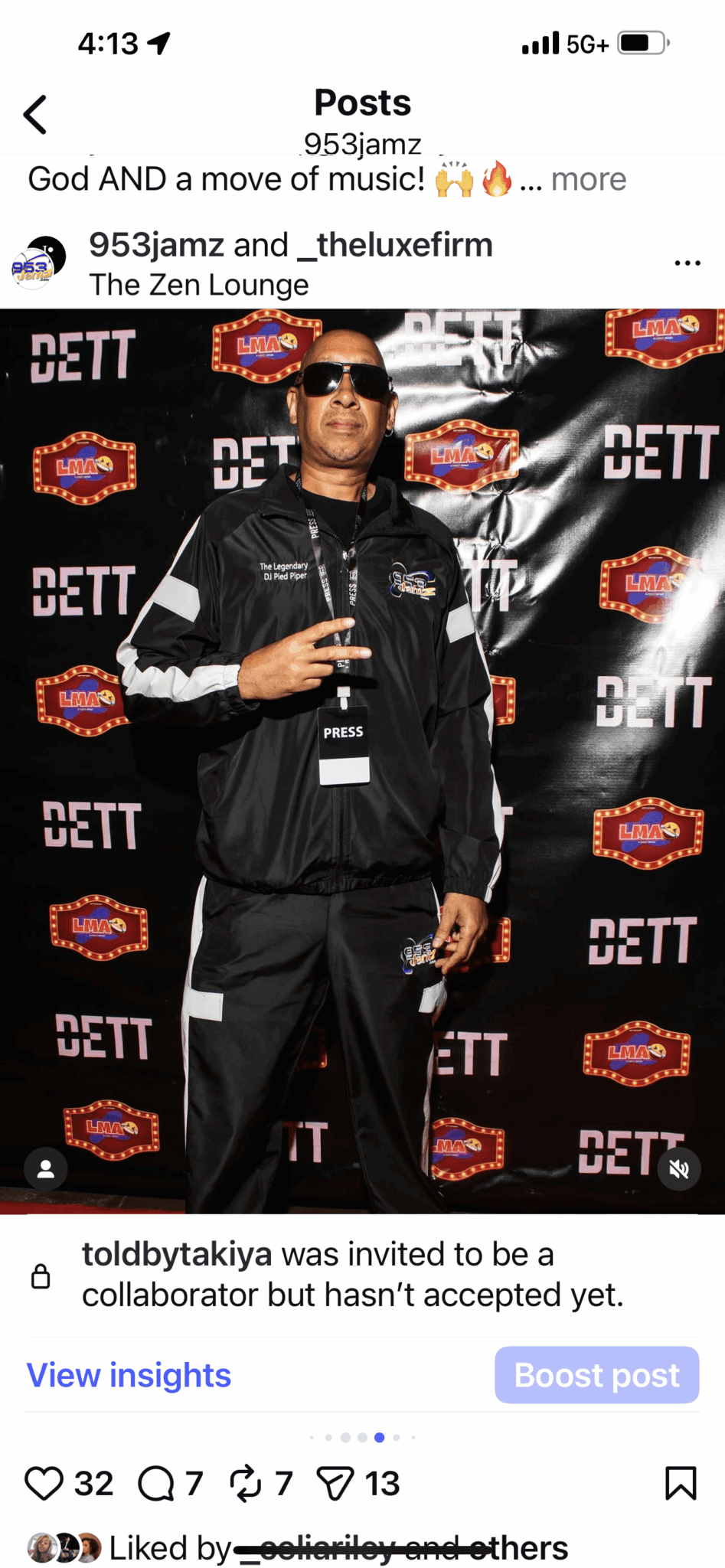 Person wearing sunglasses and a black jacket holding a drink, standing in front of a black backdrop with logos.