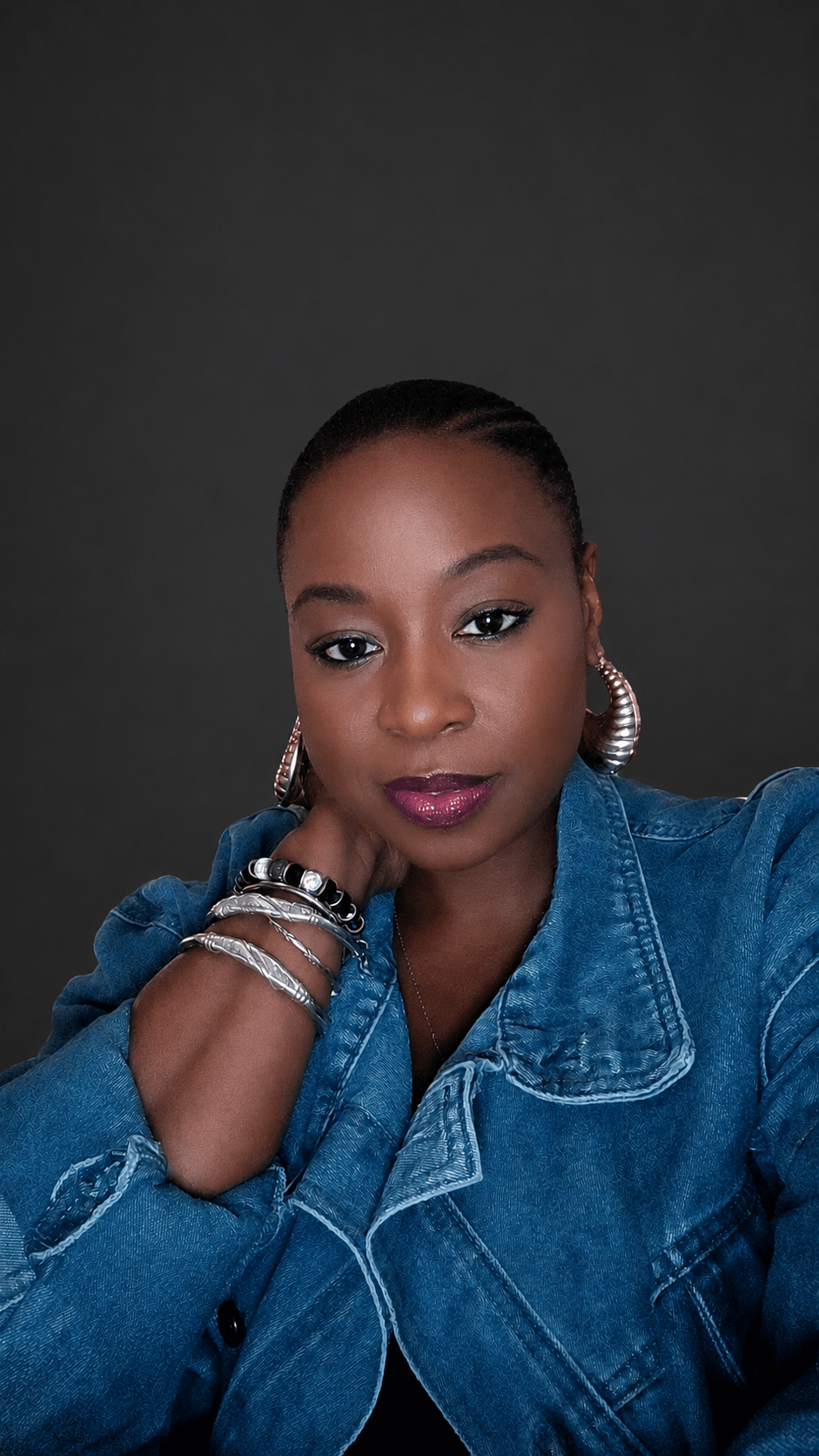Portrait of a woman with dark hair, wearing a denim jacket and large earrings, against a dark background.