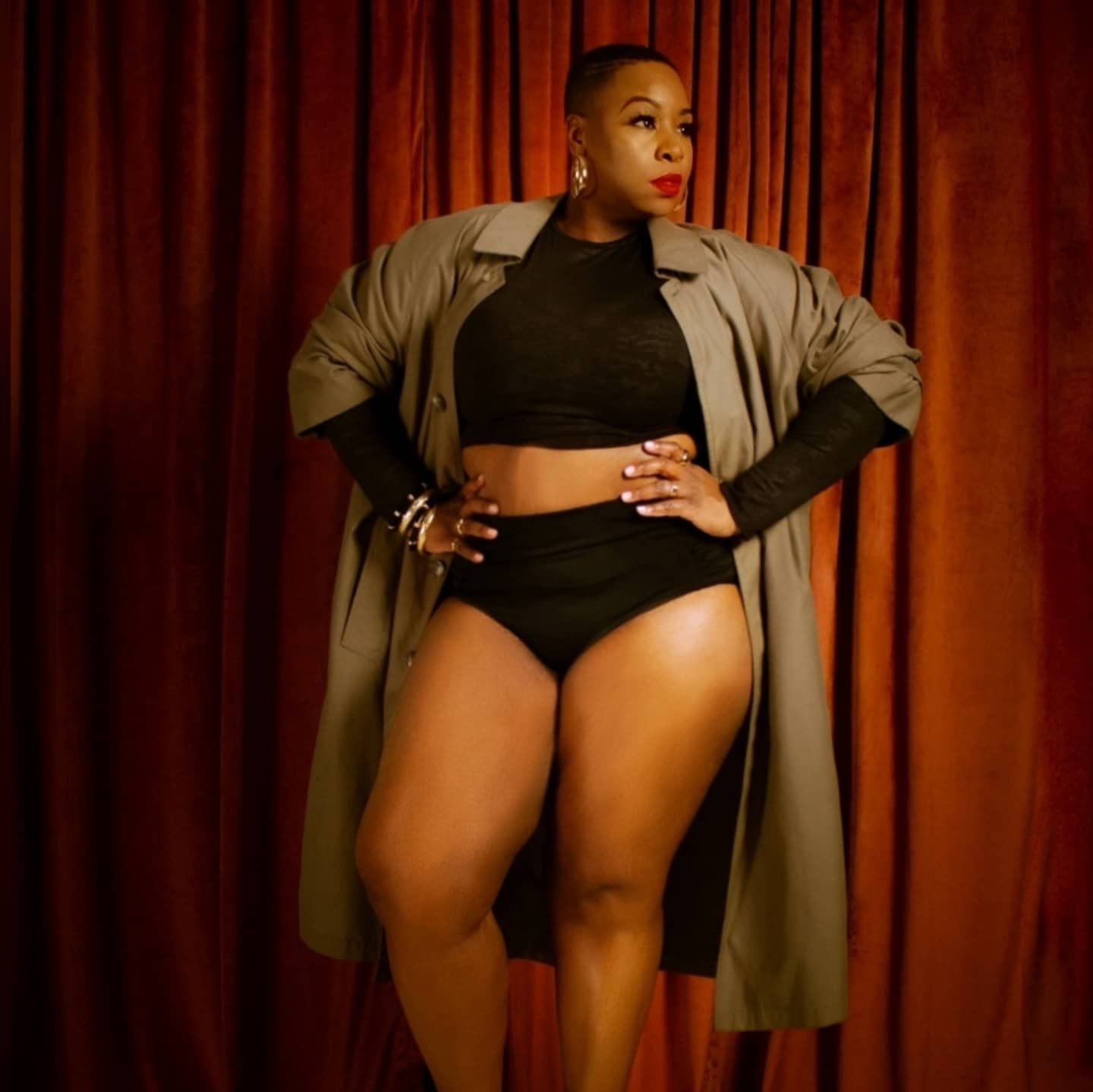 Woman with short hair and bold makeup stands with hands on hips in front of red curtains, wearing a black crop top and shorts, with a long coat.