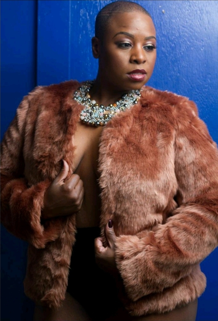 Woman with short hair wearing a fur coat and a large pearl necklace, standing against a blue wall.