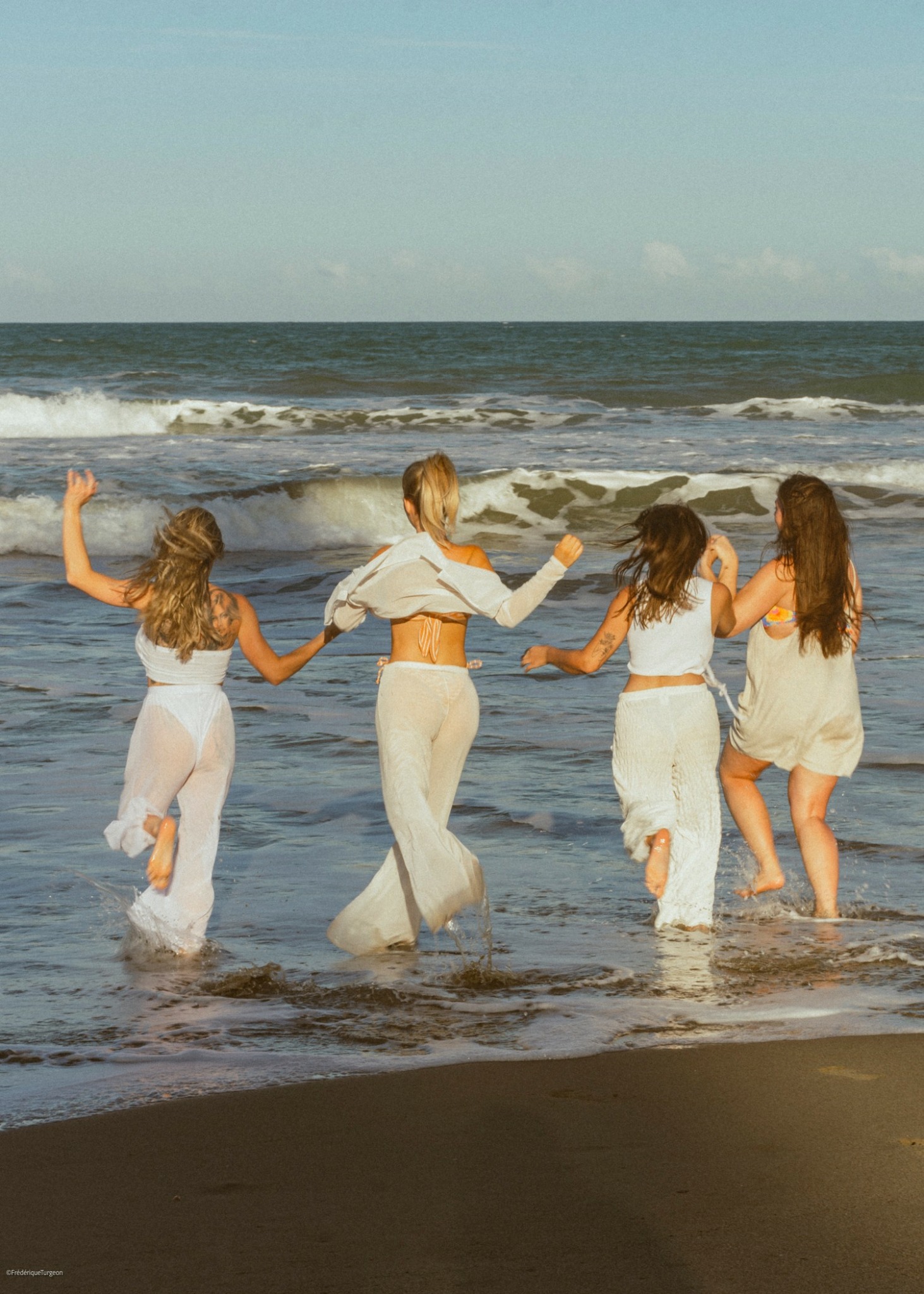Four girls holding hands and walking in the ocean waves at the beach, with the sky and sea in the background.