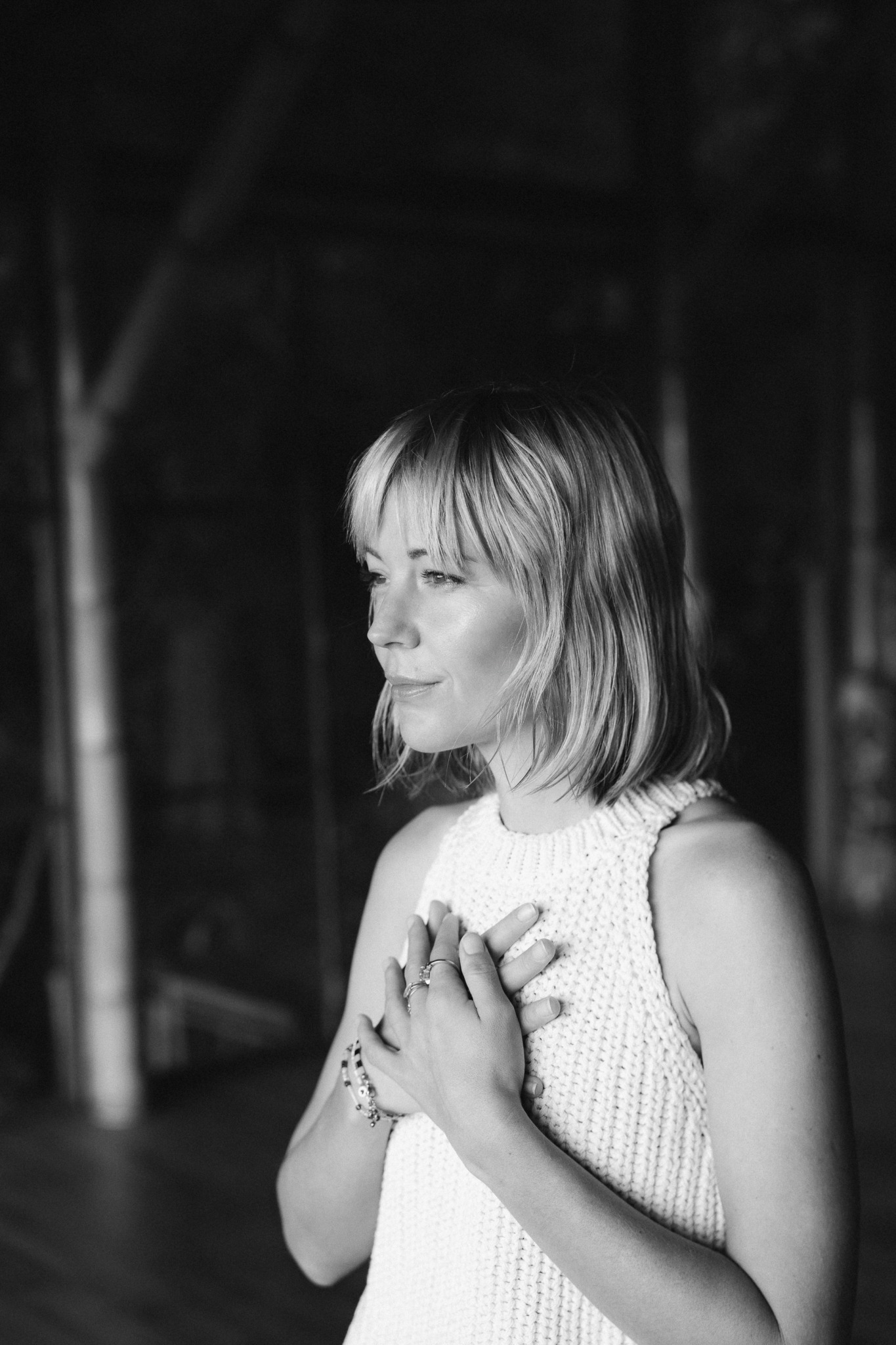 Young woman with shoulder-length hair in a sleeveless top, hands on chest, looking to the side, in a dark setting.