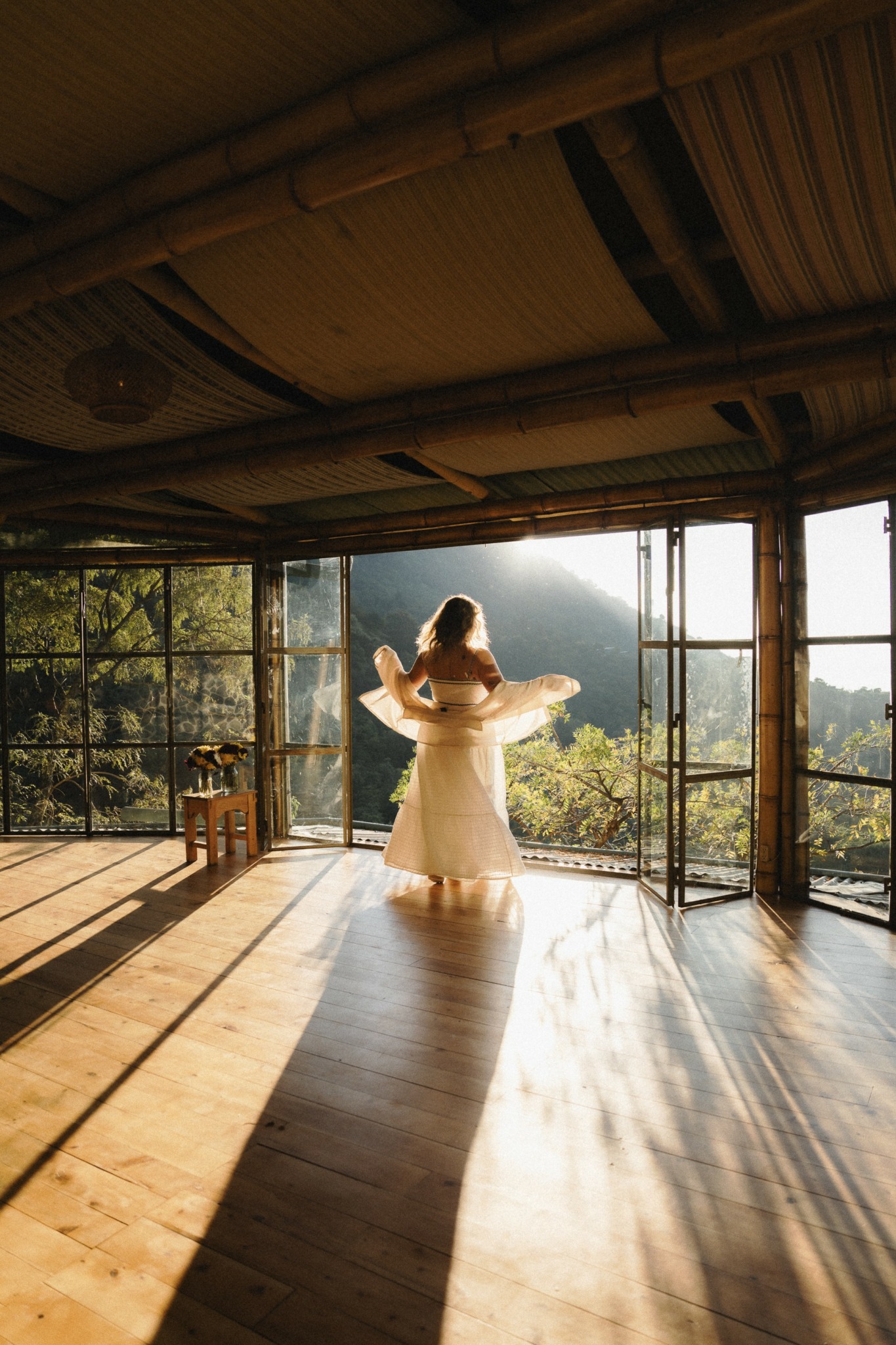 Person in a white dress standing on a wooden floor, looking out through large open windows at a scenic landscape, sunlight streaming in.