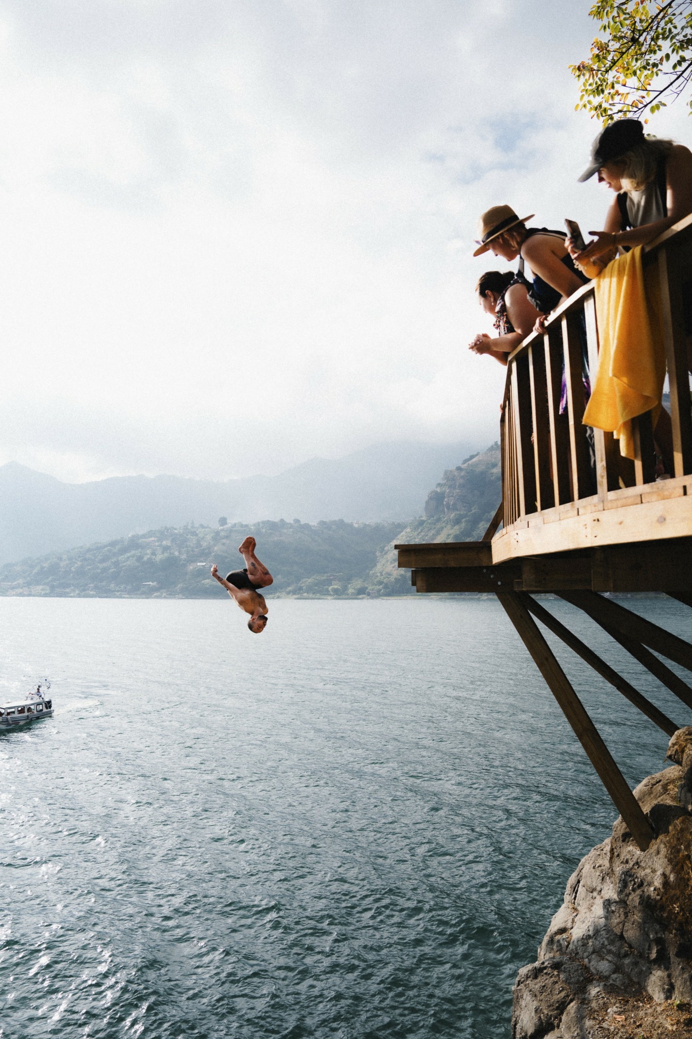 People on a wooden deck overlooking a lake, with one person jumping into the water, mountains in the background.