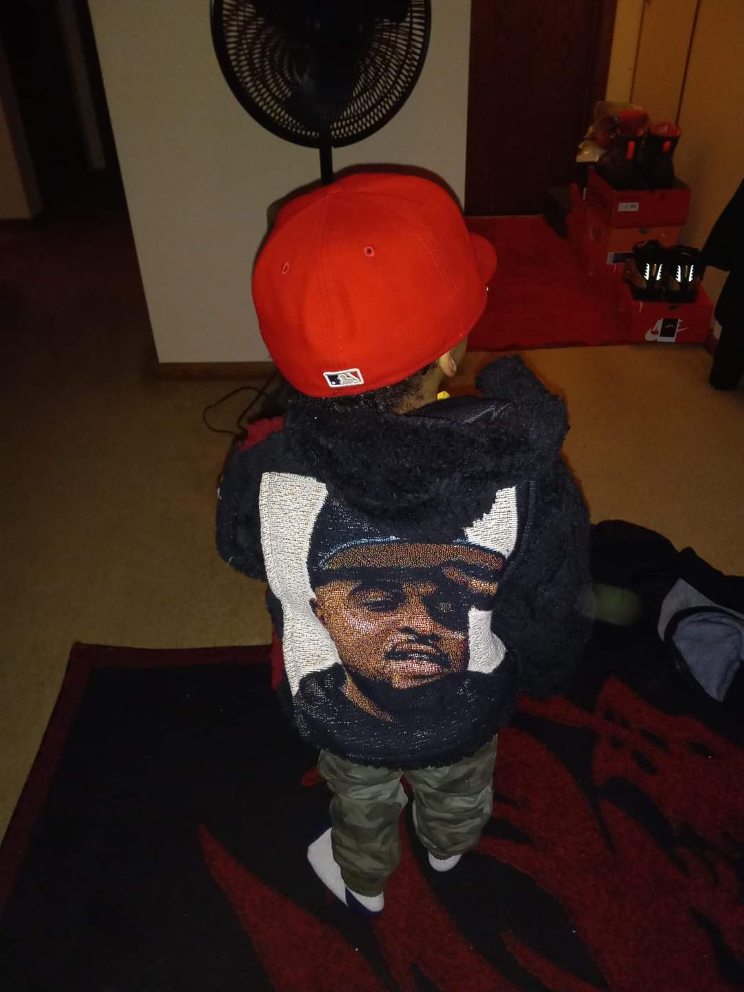 Child wearing a red cap and a hoodie with a large portrait of a man on the back, standing indoors near a fan.