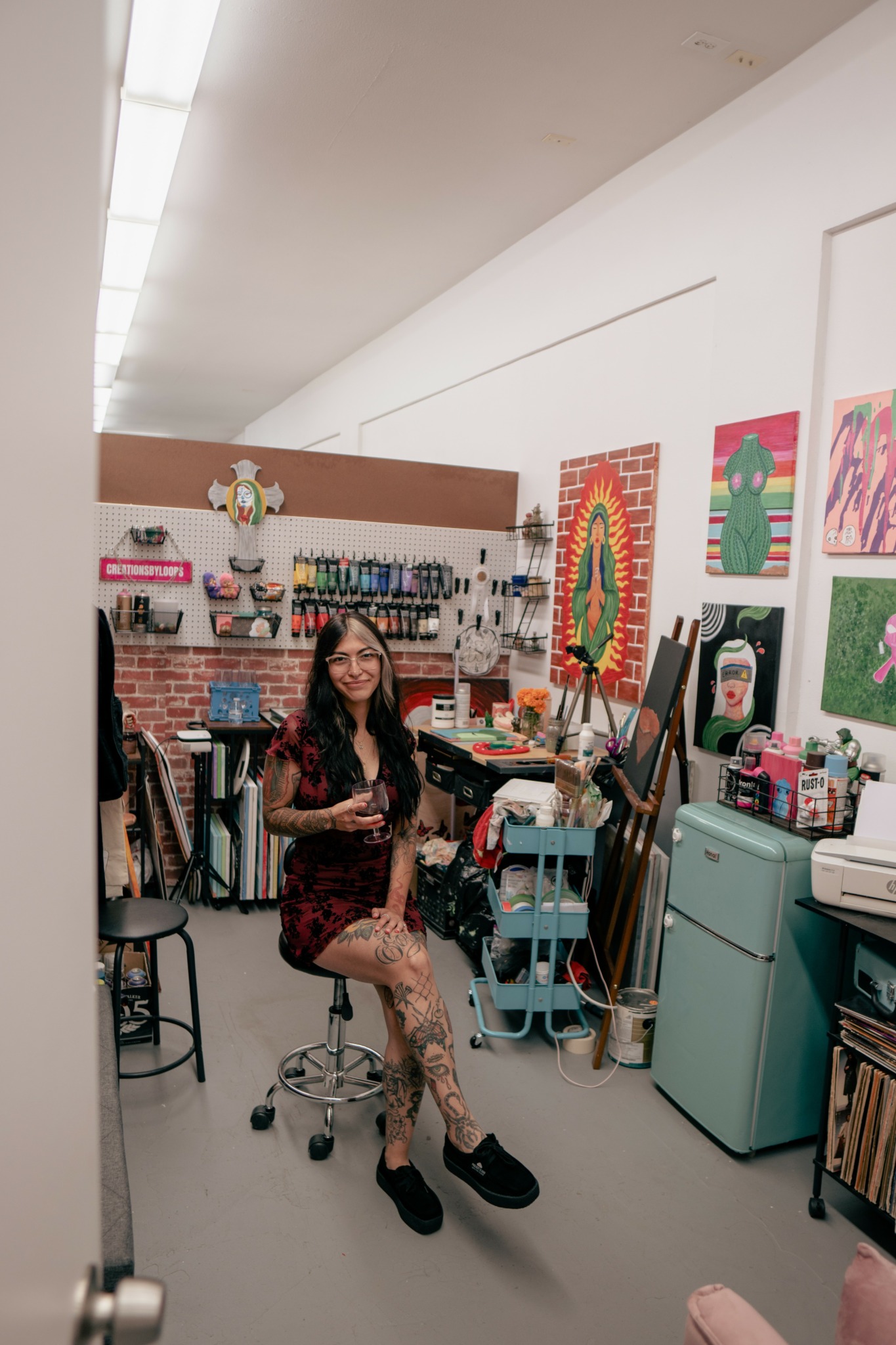 Woman sitting on a stool in a colorful art studio with paintings and supplies, smiling at the camera.
