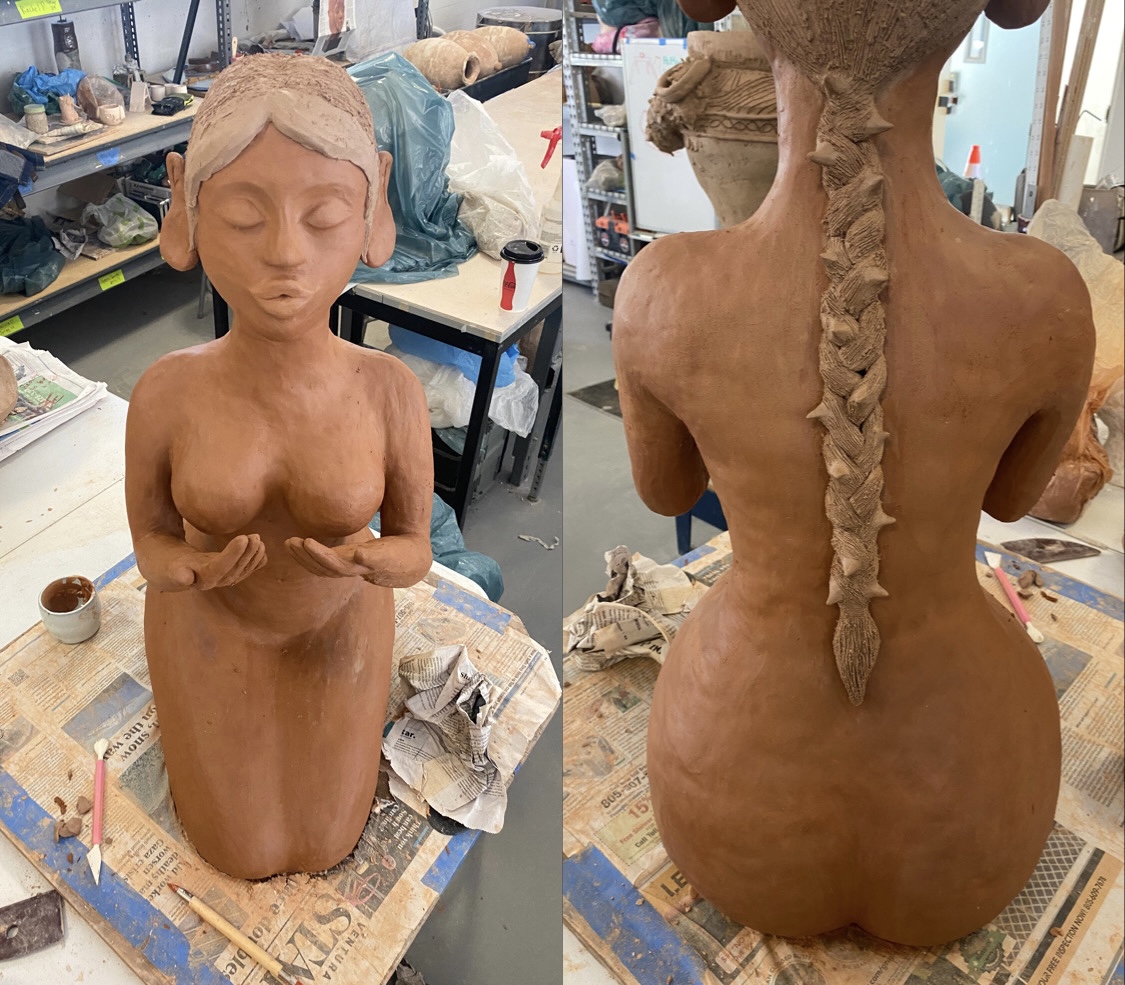 Unfinished clay sculpture of a woman with long braided hair, seen from the back, in a workshop setting.