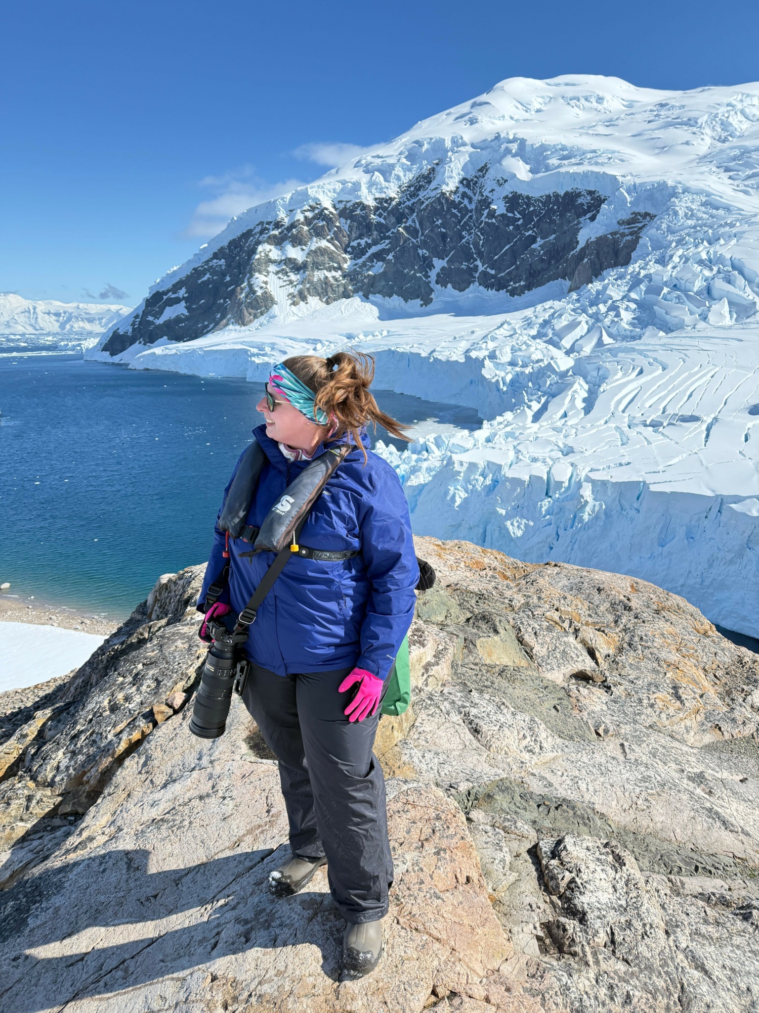 Person standing on rocky shore with snow-capped mountain and blue sky in background.
