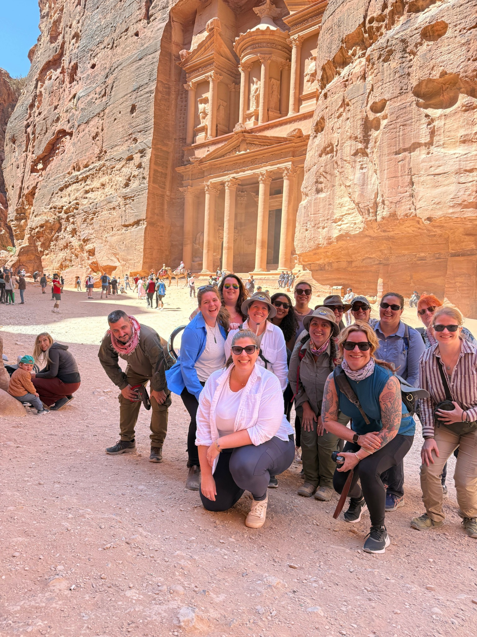Group of people standing and sitting outdoors near a rocky cliff, some smiling and looking at the camera.