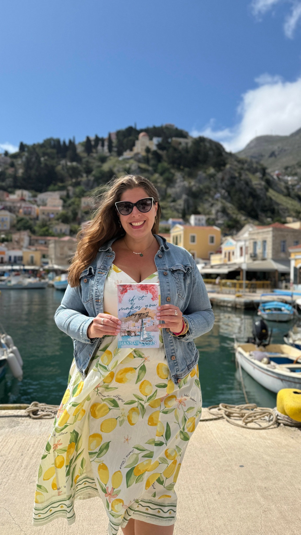 Woman in sunglasses holding a map, standing near boats with a hillside town in the background.