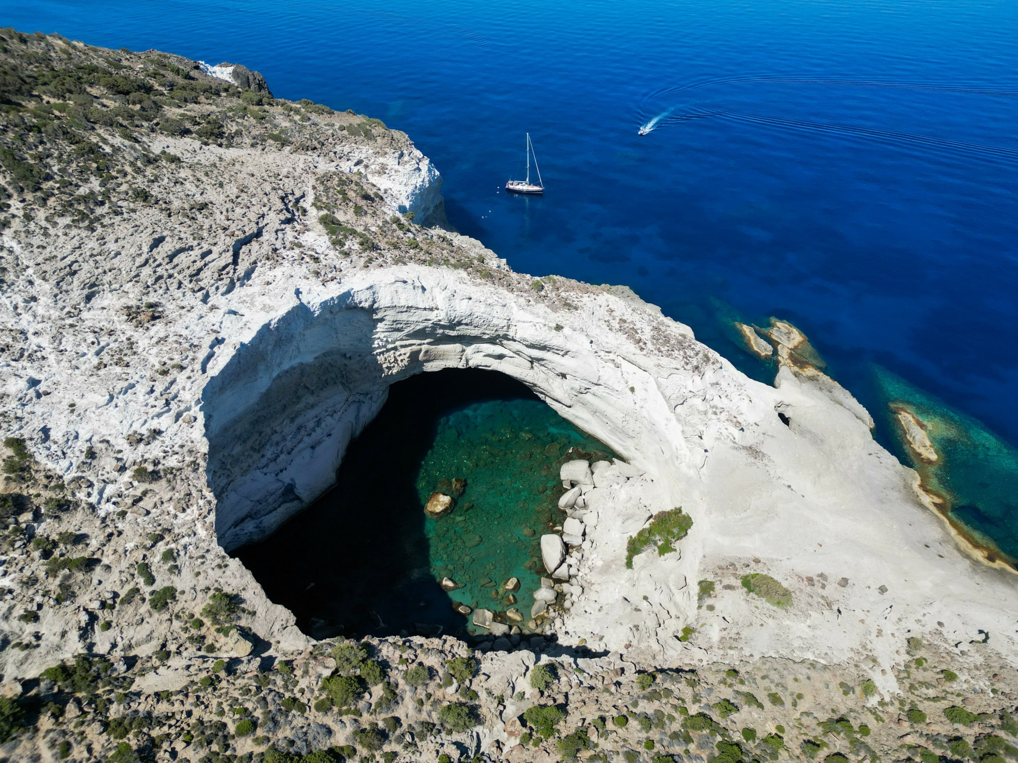 A coastal landscape with a large natural arch formation over a small water body, and a sailboat in the sea.
