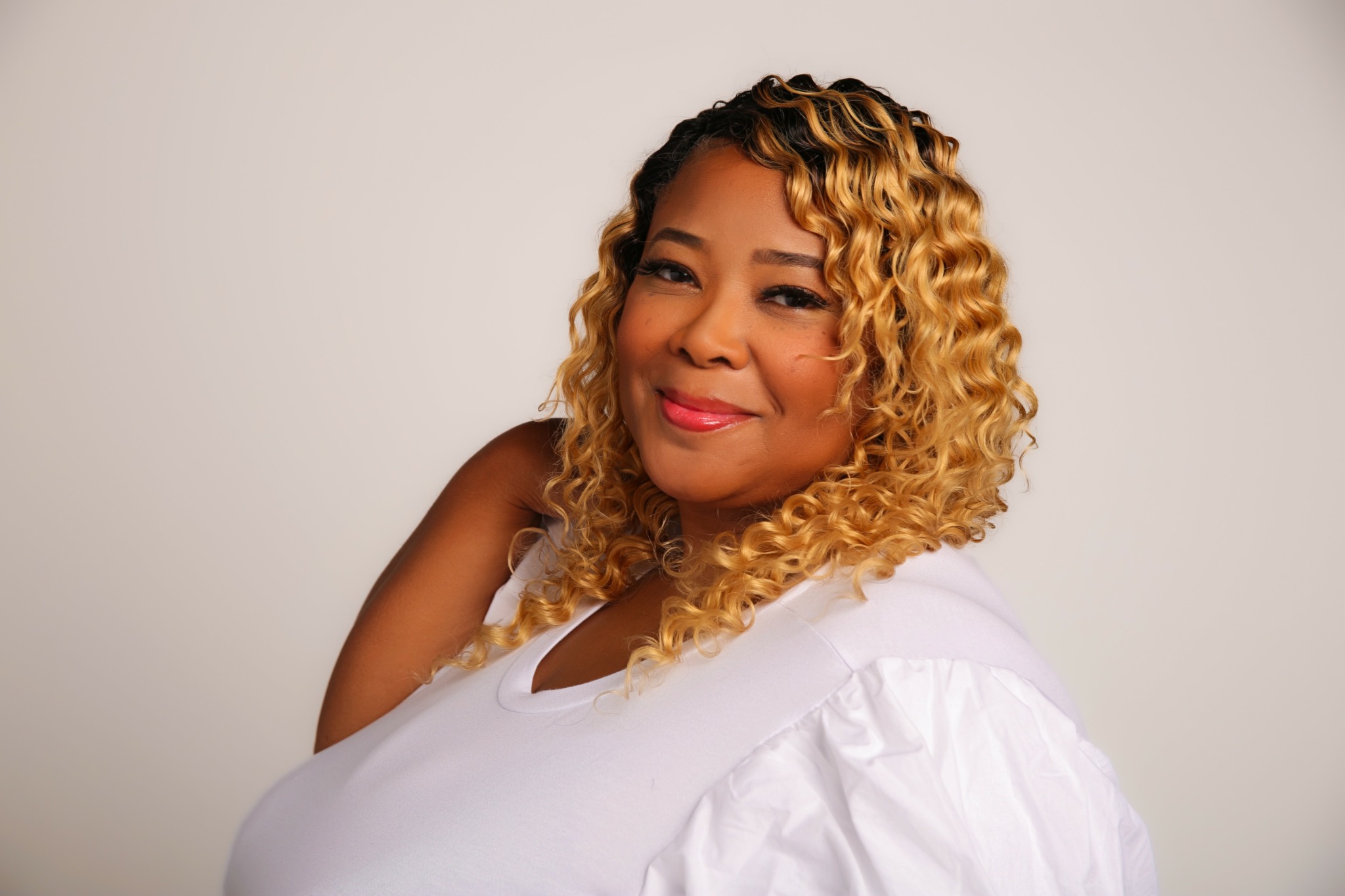 Woman with curly blonde hair smiling, wearing a white top, against a plain background.