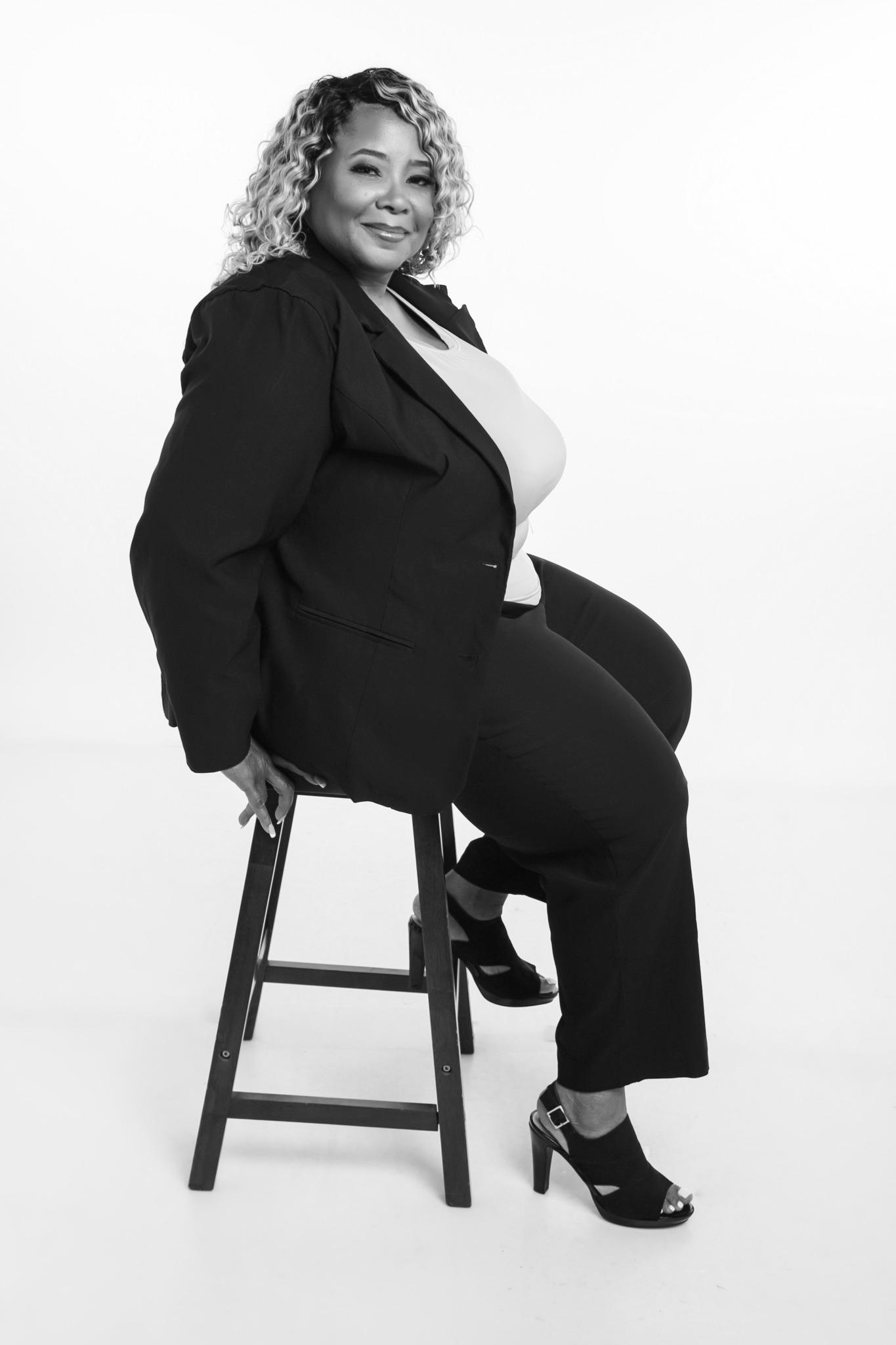 Woman sitting on a stool, smiling, wearing a blazer and high heels, in black and white.