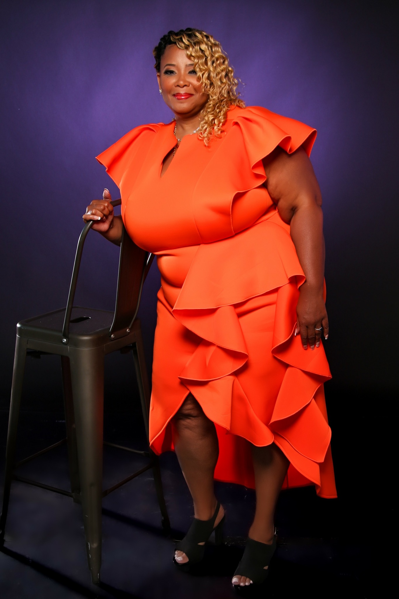 Woman in bright orange dress with ruffles, standing next to a black chair, smiling, dark background.