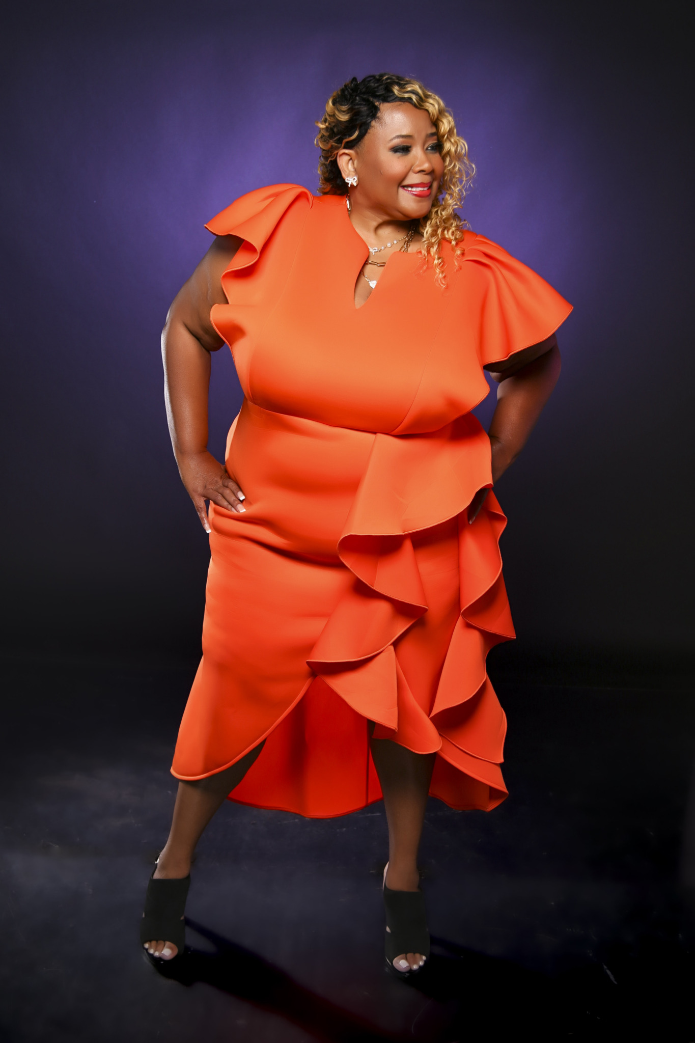 Woman in bright orange dress with ruffles, smiling, standing against a dark background.