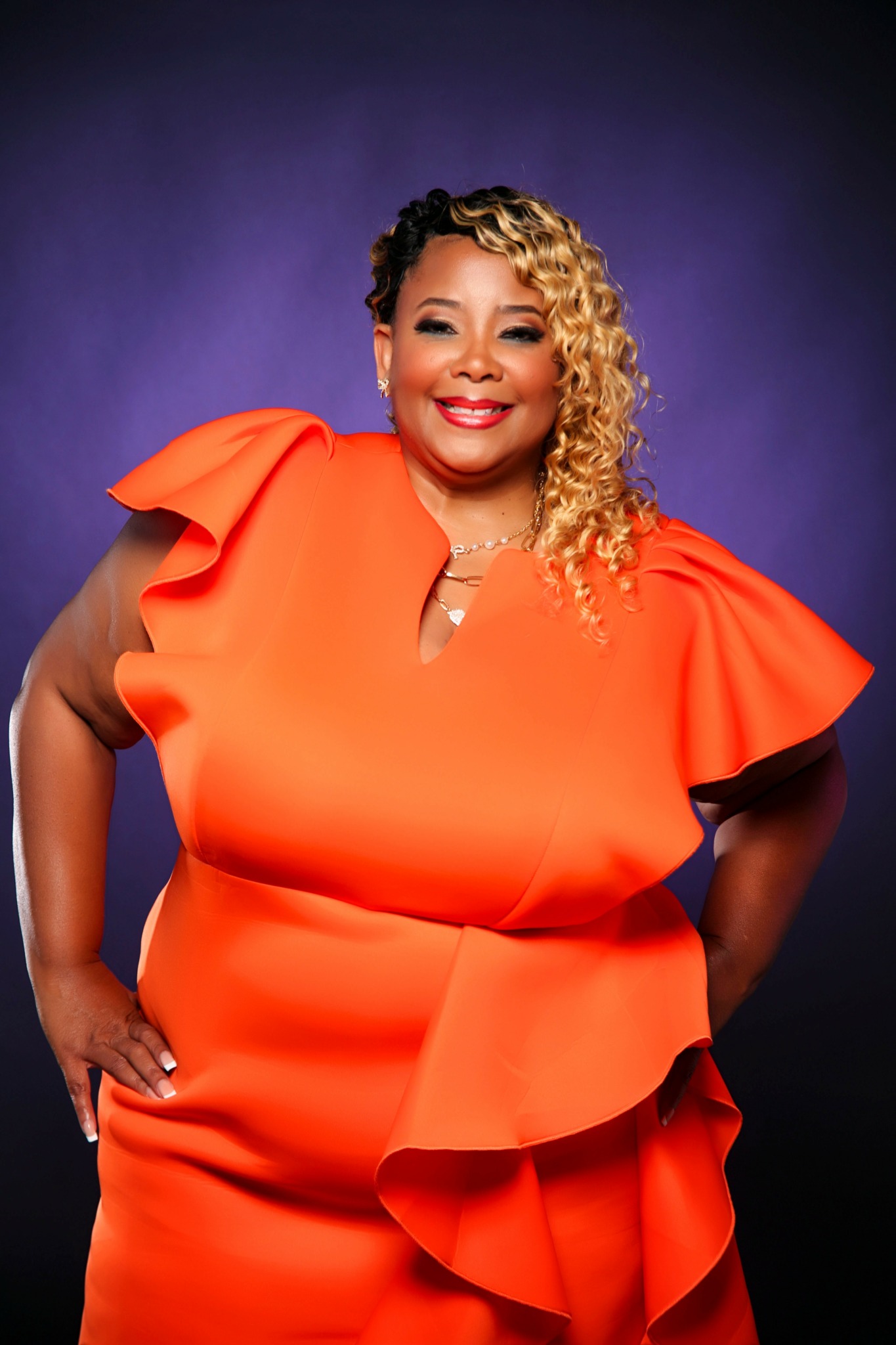 Woman with curly blonde hair wearing an orange dress with ruffled sleeves, smiling, against a purple background.