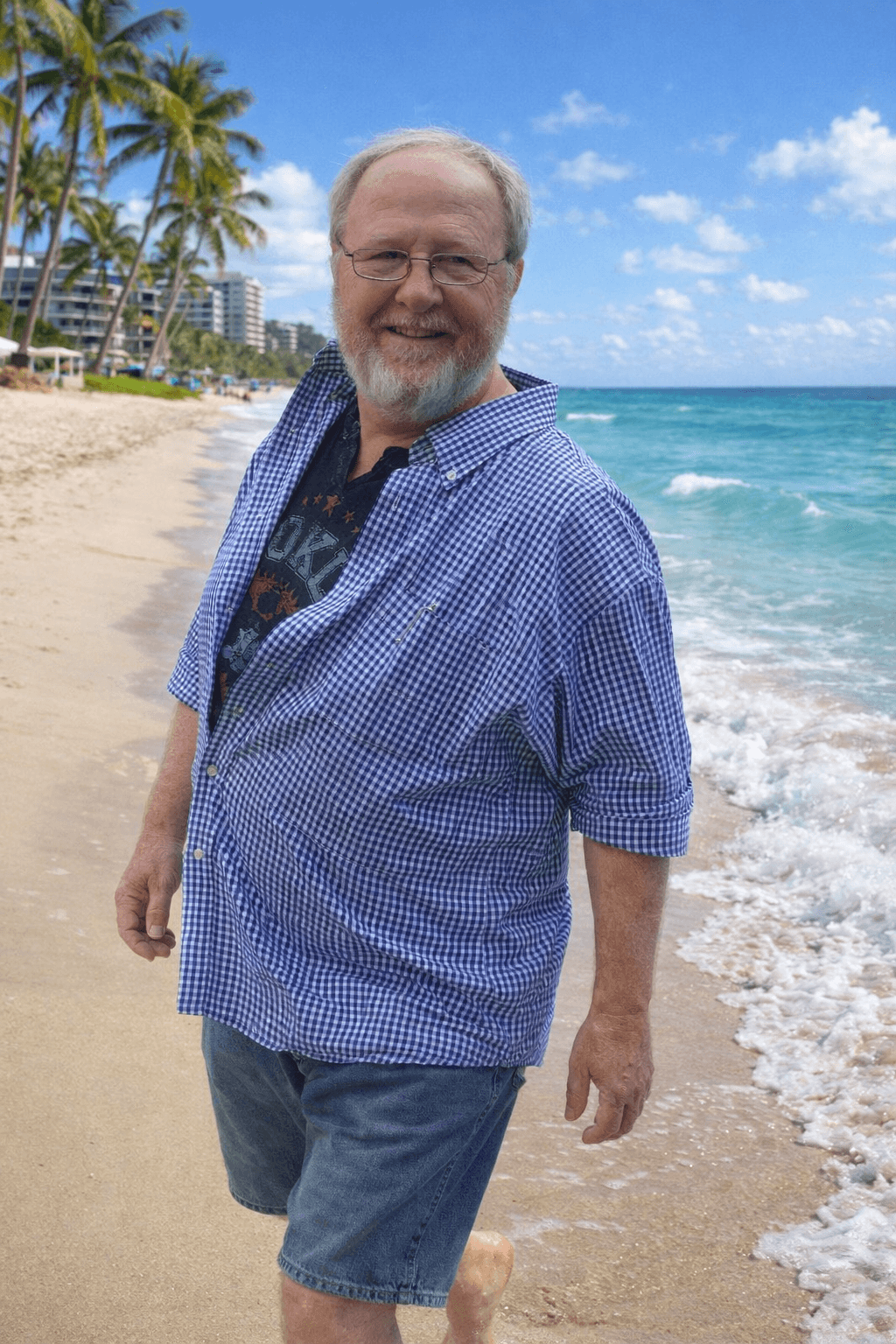 Man with glasses and beard smiling on beach, ocean and palm trees in background.