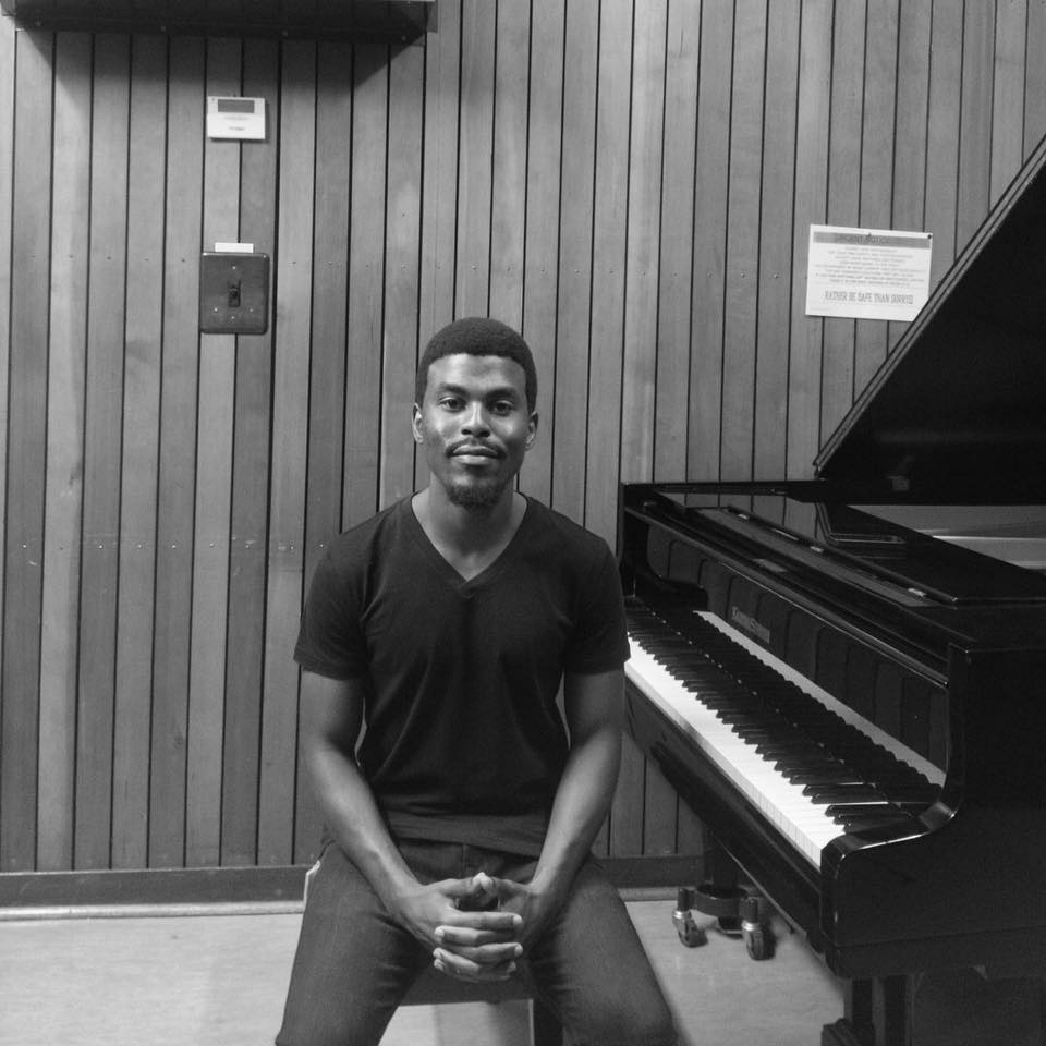 Young man sitting with hands clasped in front of a piano in a room with wooden panel walls.