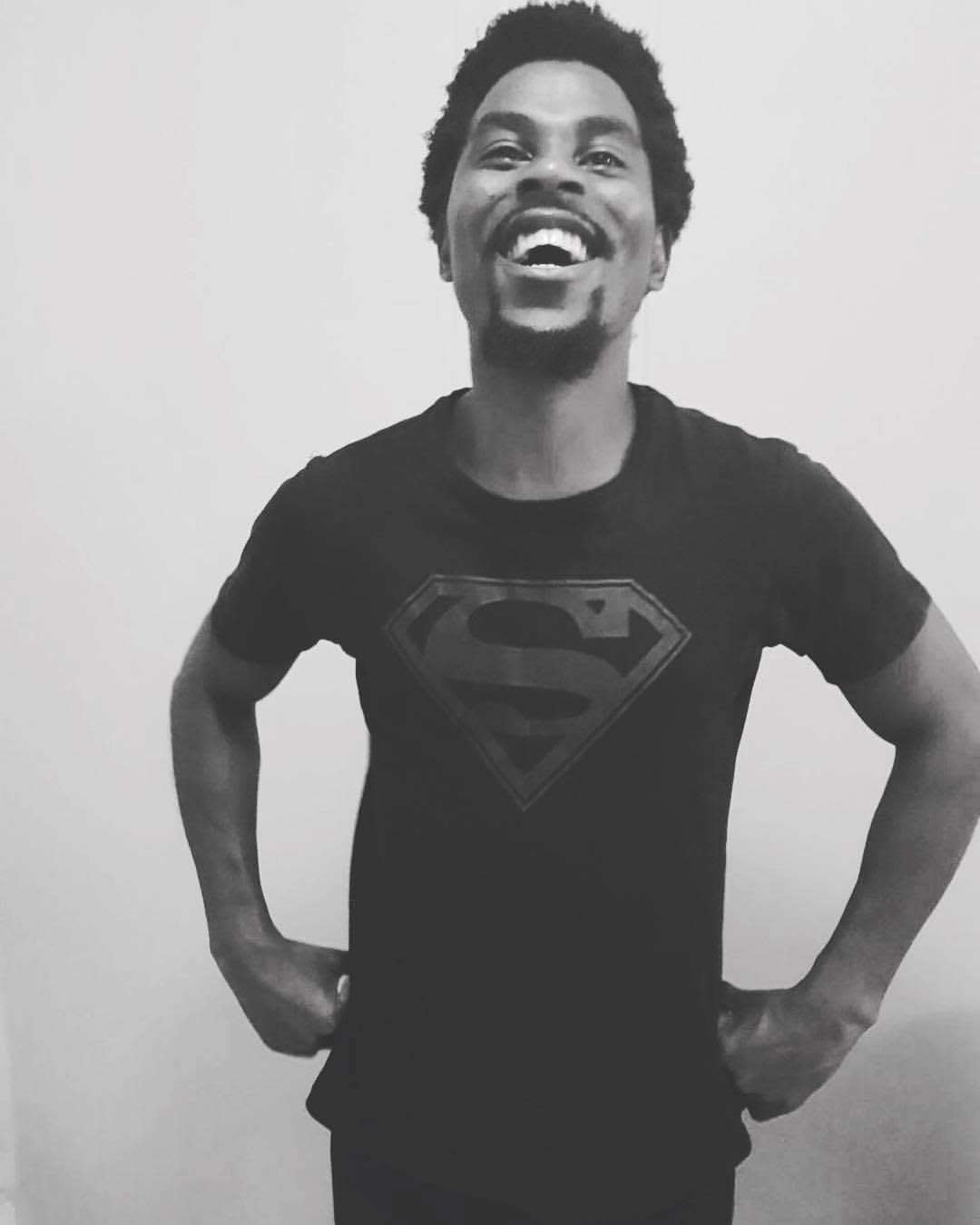 Person with short curly hair smiling, hands on hips, wearing a Superman logo T-shirt, standing against a plain background.