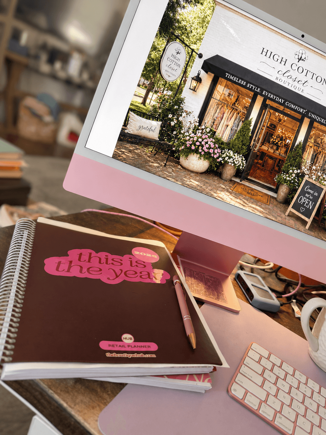 Computer monitor displaying a storefront with flowers outside, pink desk with a planner, pen, and keyboard, and a mug.