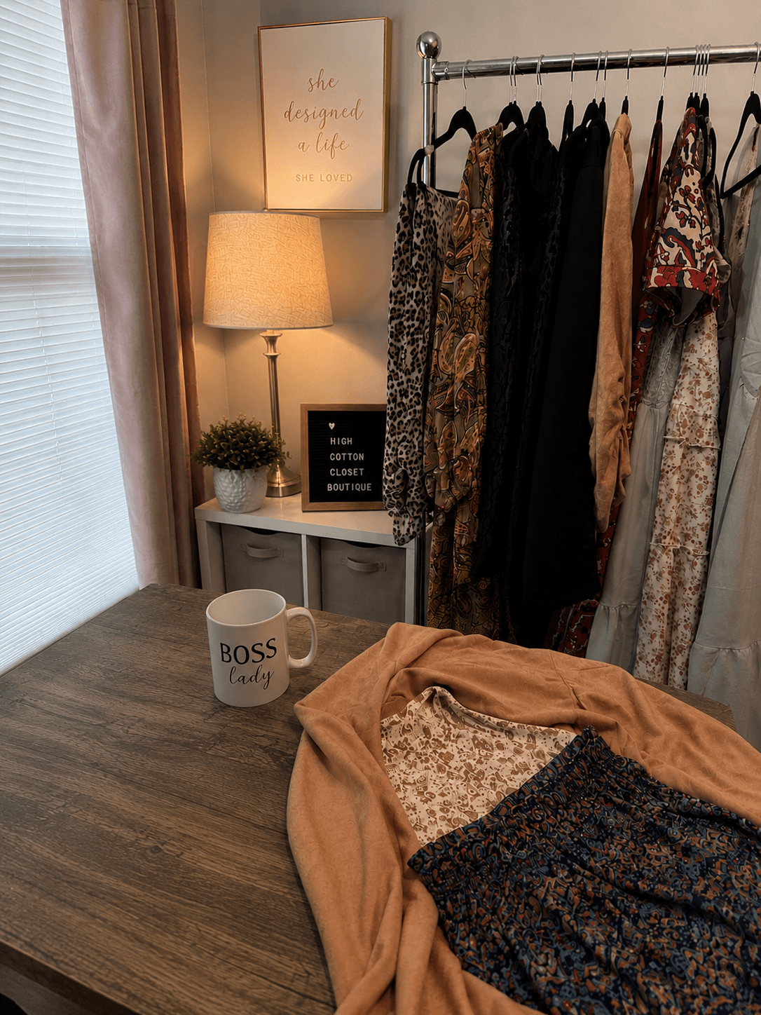 Clothing rack with various dresses and garments, side table with lamp, plant, and BOSS mug, and a bed with clothes, in a cozy room.