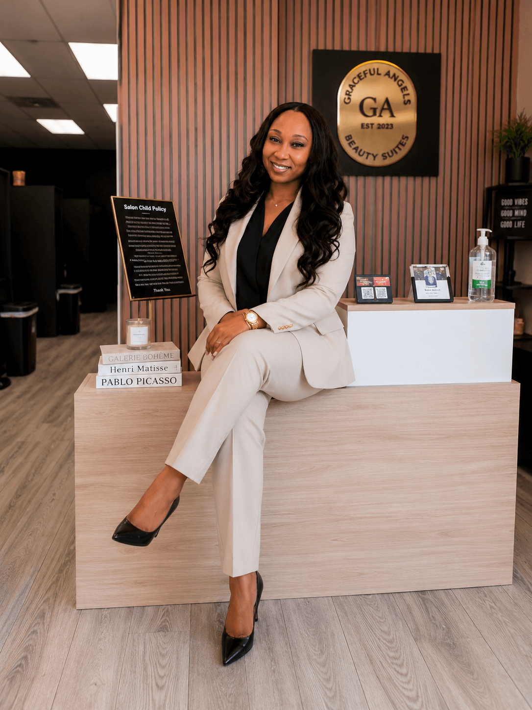 Woman in white suit sitting on reception desk, smiling, in office with wood panel wall and GA logo, hand on desk, black shoes